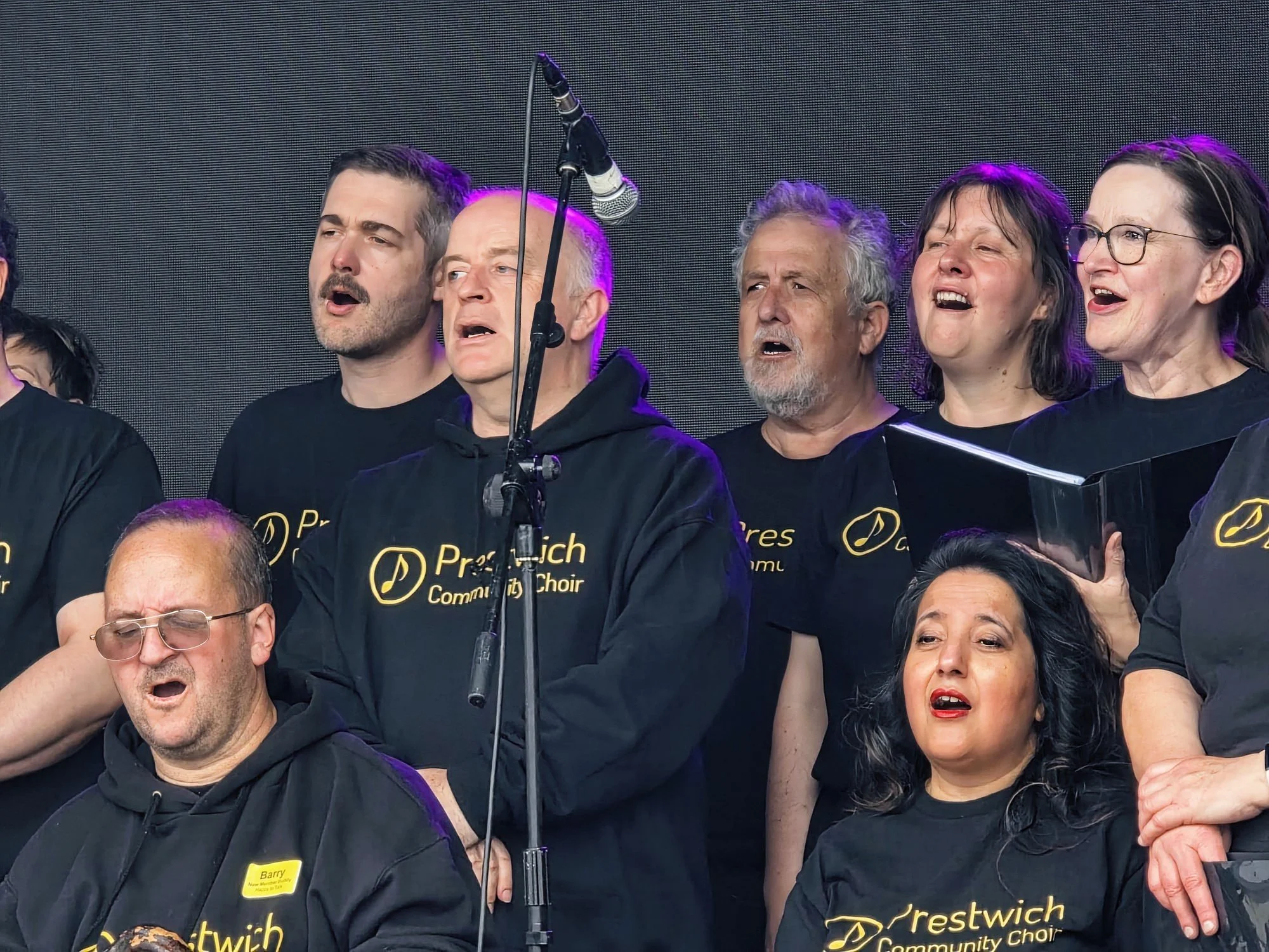 A choir performing on stage, with members wearing black T-shirts that say 'Prestonch Community Choir' in yellow text, and singing with expressions of concentration and emotion.