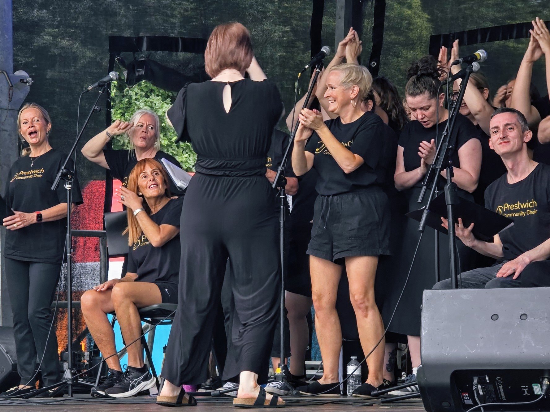 A group of people on stage, part of a choir, wearing black shirts with 'Prestwich Community Choir' written on them. Some are standing and singing, while one woman is seated, surrounded by others who are clapping and engaging with a conductor or speak
