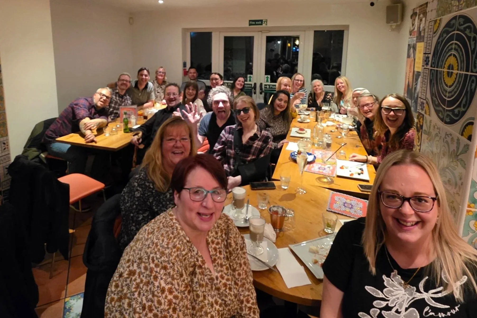 A group of people gathered around a long wooden table in a restaurant, smiling and enjoying drinks and food, celebrating together.