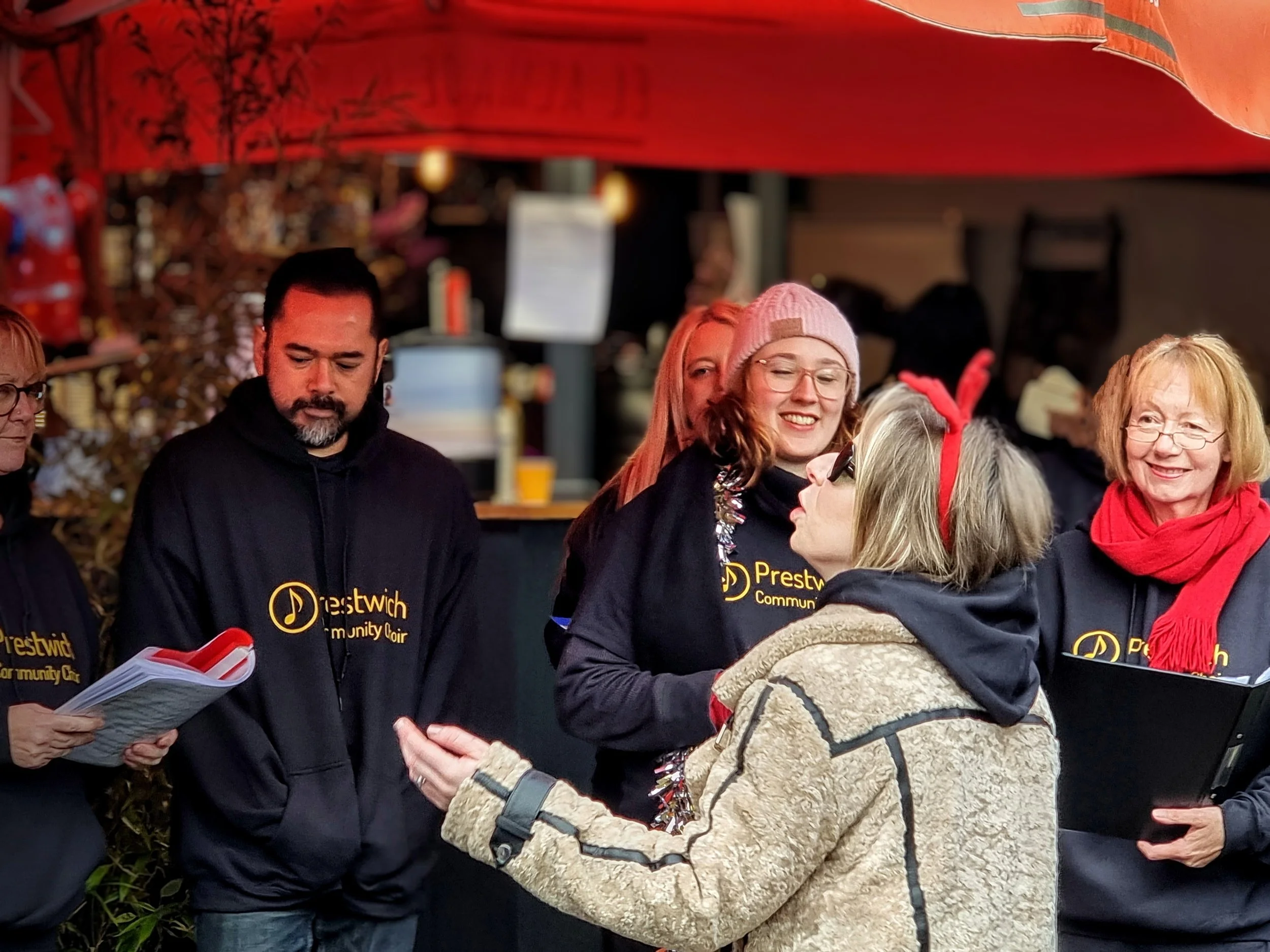 A group of people, wearing black hoodies with yellow text, are gathered outdoors under a red canopy, engaging in conversation and holding papers, with some smiling and interacting.