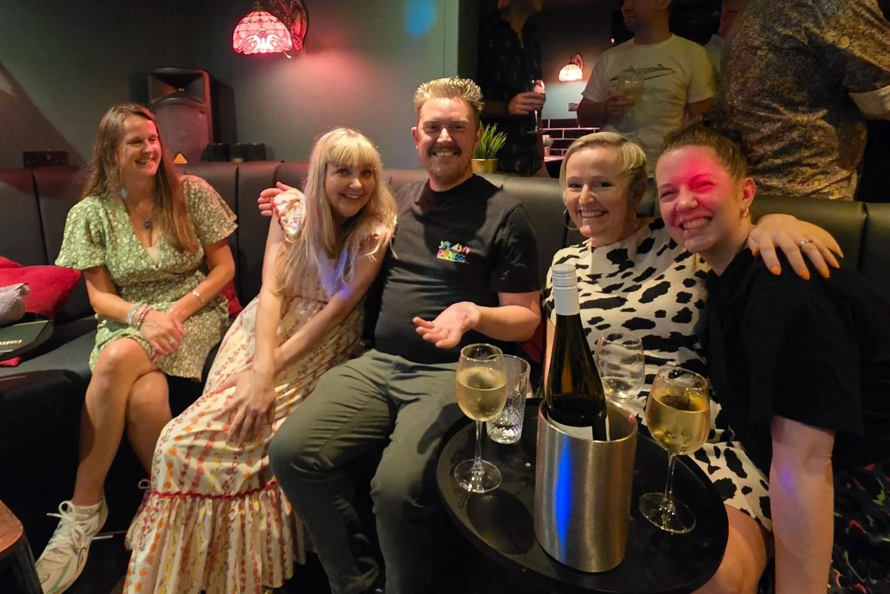 Group of five friends enjoying drinks at a bar, sitting closely on a black leather couch, smiling, with some holding glasses of wine. There are two women and three men, one man with with gray hair, all appearing to have a good time in a dimly lit setting with red hanging lamps.