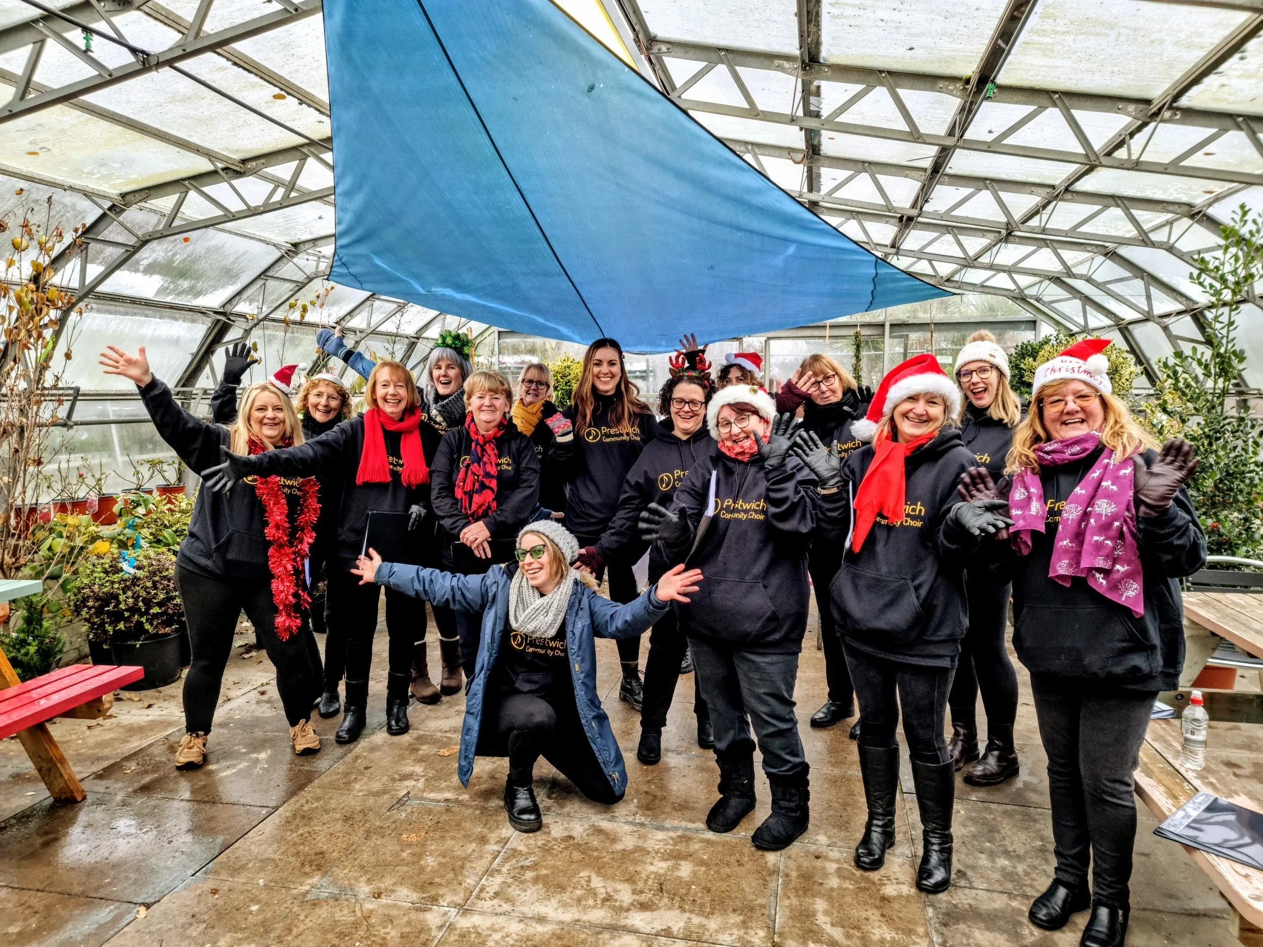 A group of choir members are dressed in Christmas hats and scarves, posing cheerfully inside a greenhouse with plants around them, celebrating the Christmas season.