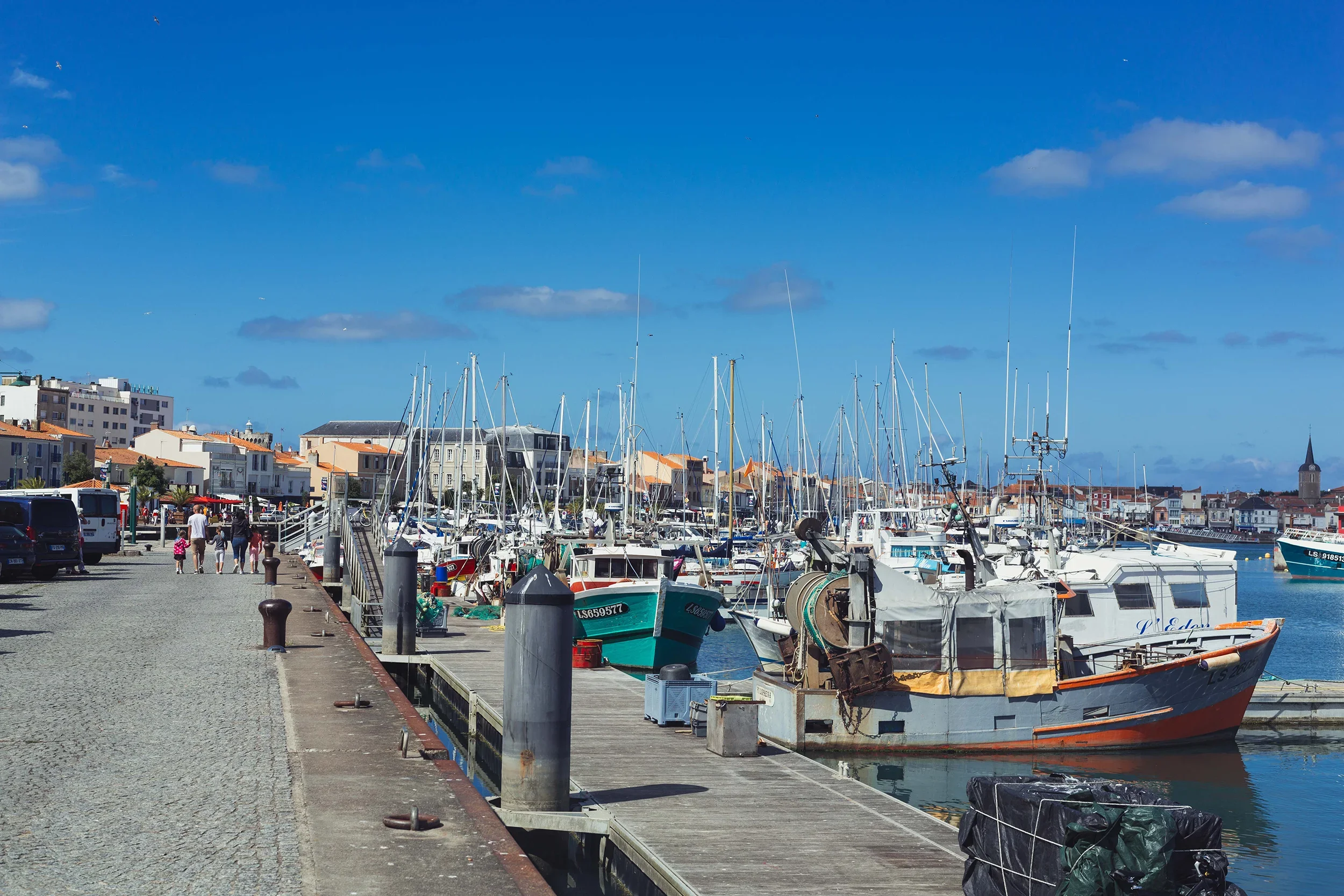 Marina with numerous boats docked, beside a pedestrian walkway with parked cars, under a blue sky with scattered clouds, buildings in the background, and a church steeple.