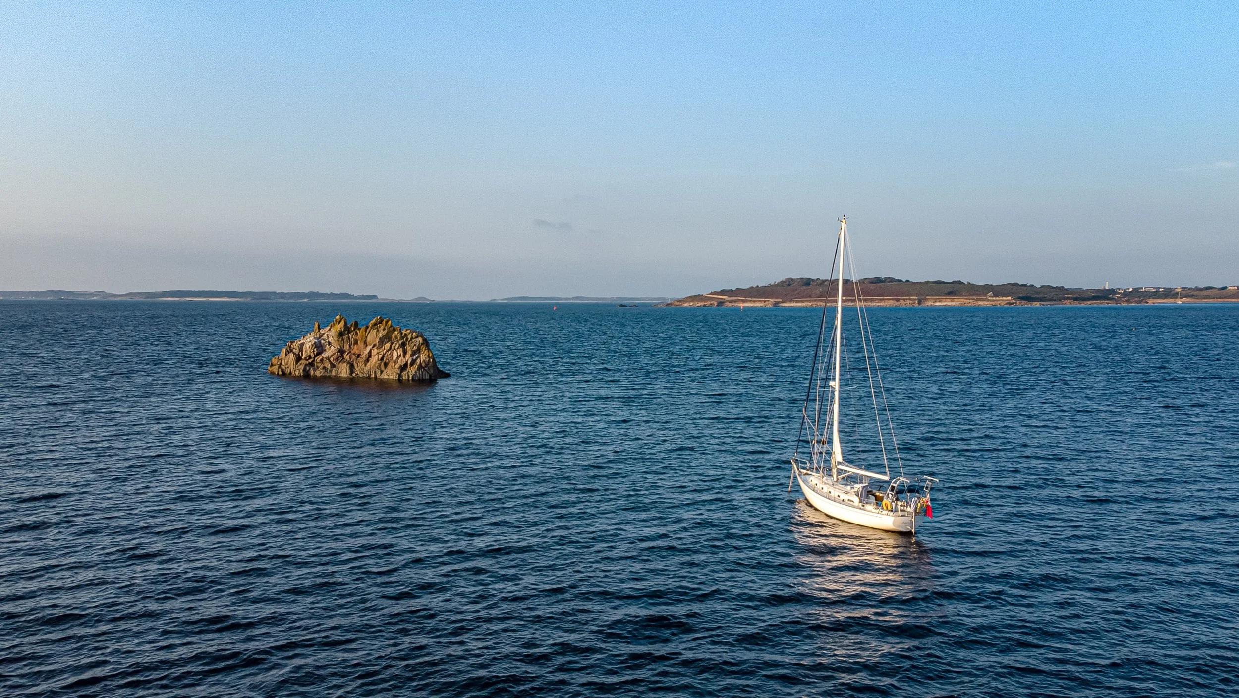 A sailboat floating on calm blue water near a small rocky island with a larger landmass in the background under a clear sky.