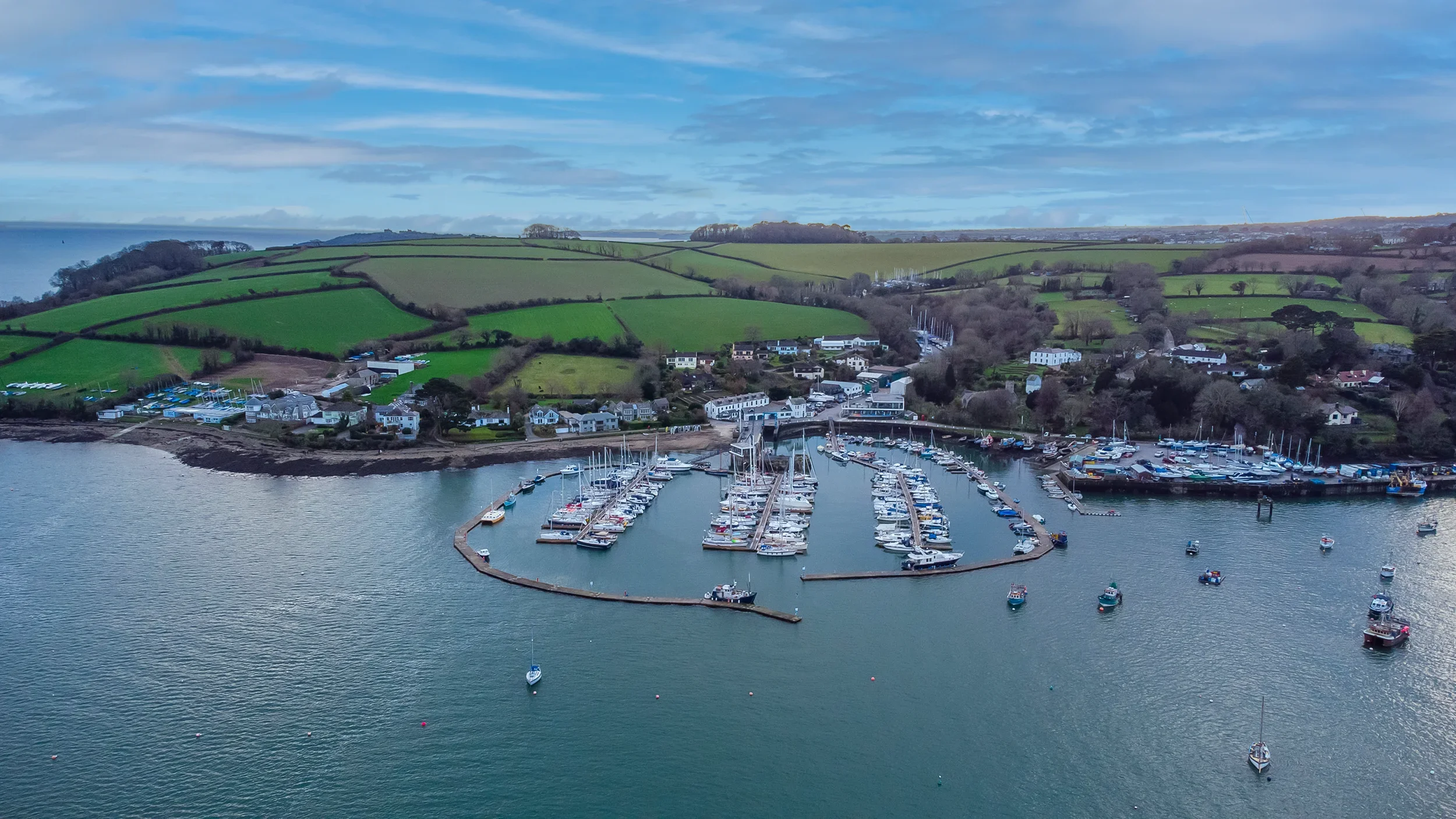 Aerial view of a marina with boats docked along piers, surrounded by green hills and scattered houses.