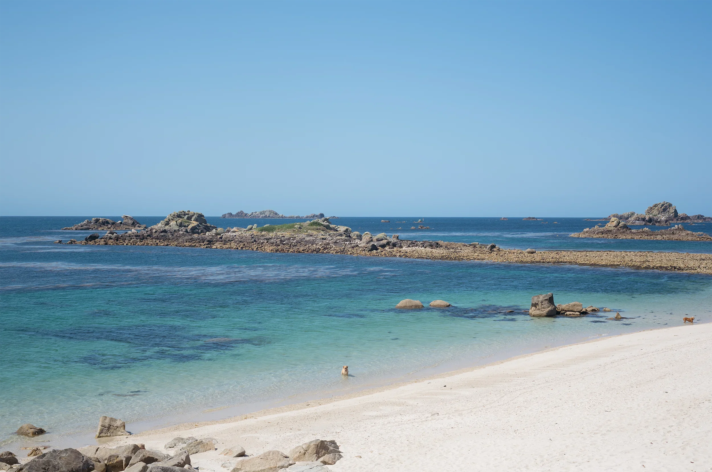 Beach scene with white sandy shoreline, rocks, turquoise water, and small islands in the distance under a clear blue sky.