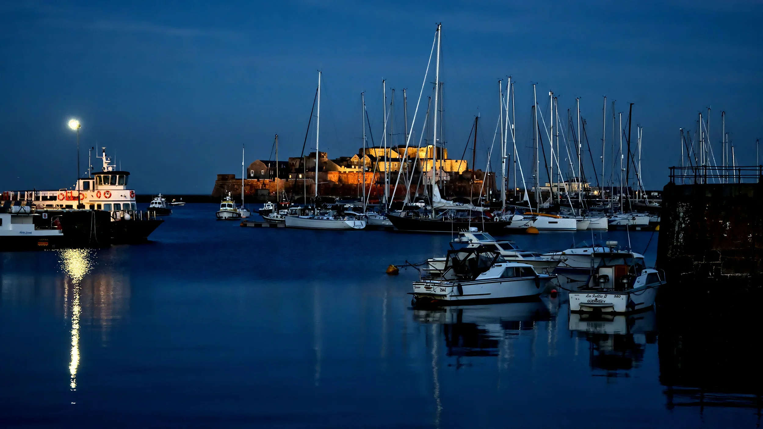 Nighttime view of a marina with boats docked, illuminated by a city light, and a historic castle in the background.