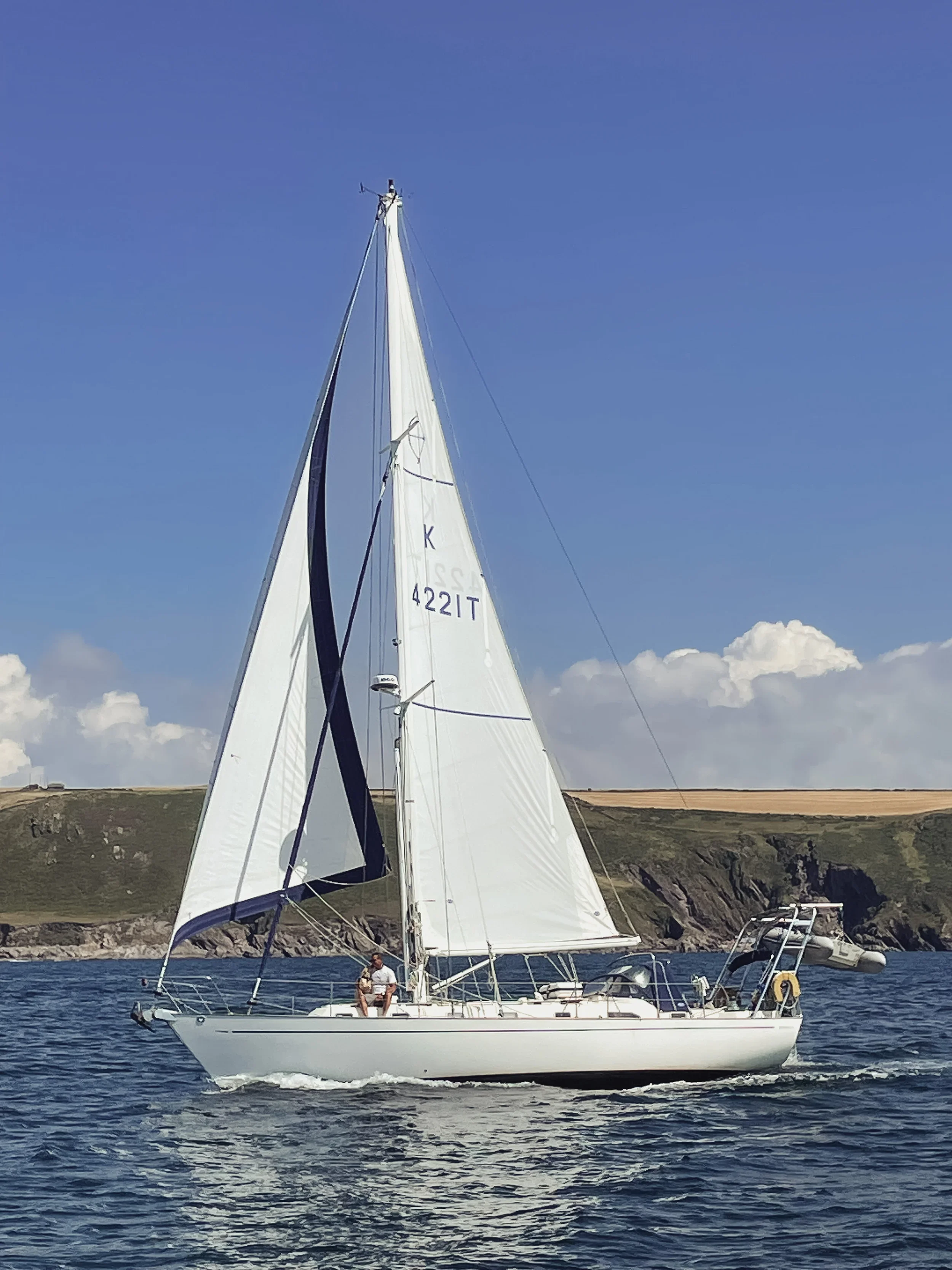 Sailboat with white sails on water, cliffs in background, clear blue sky.