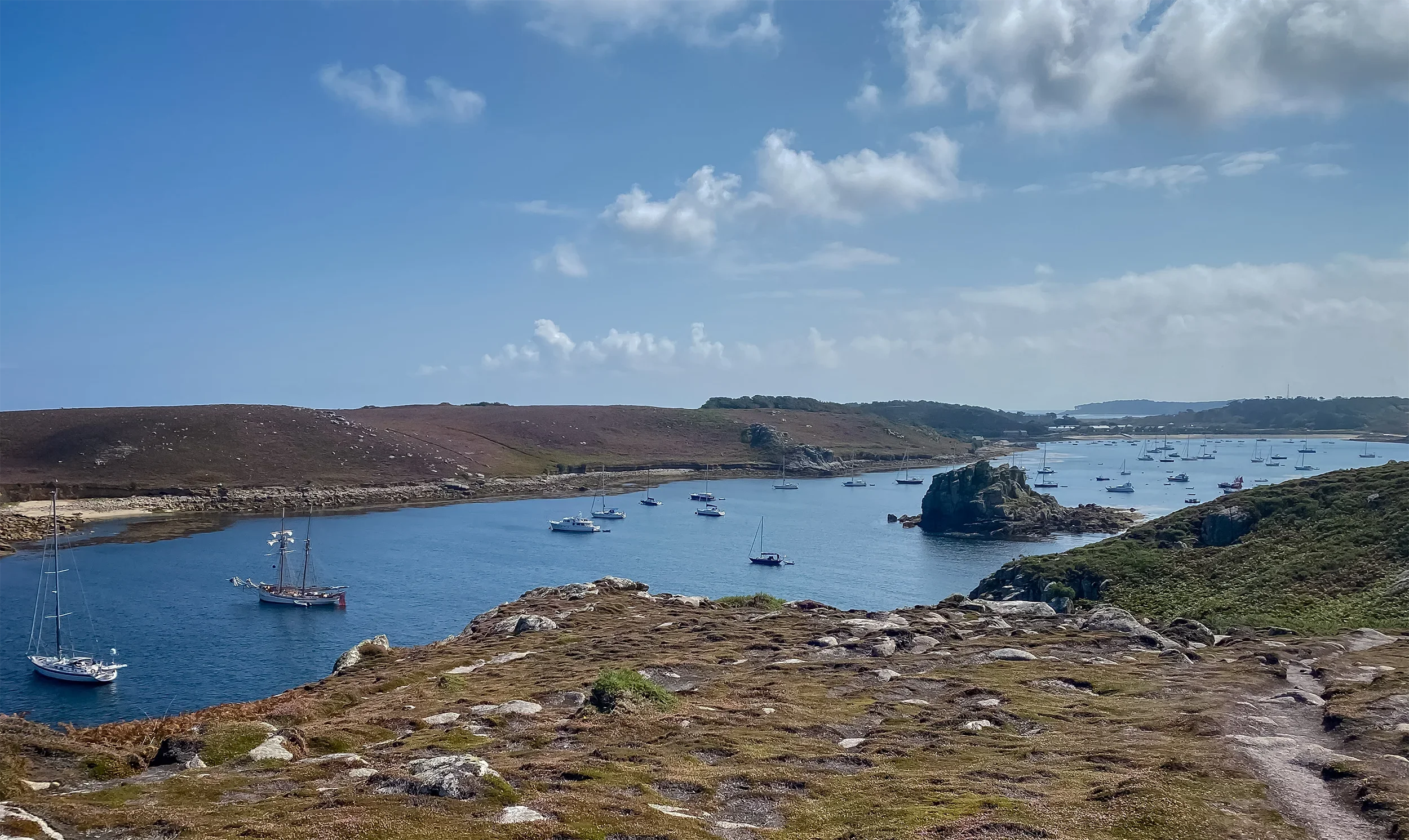 A scenic view of a harbor with sailboats anchored in calm blue water, surrounded by rolling hills and rocky terrain under a partly cloudy sky.
