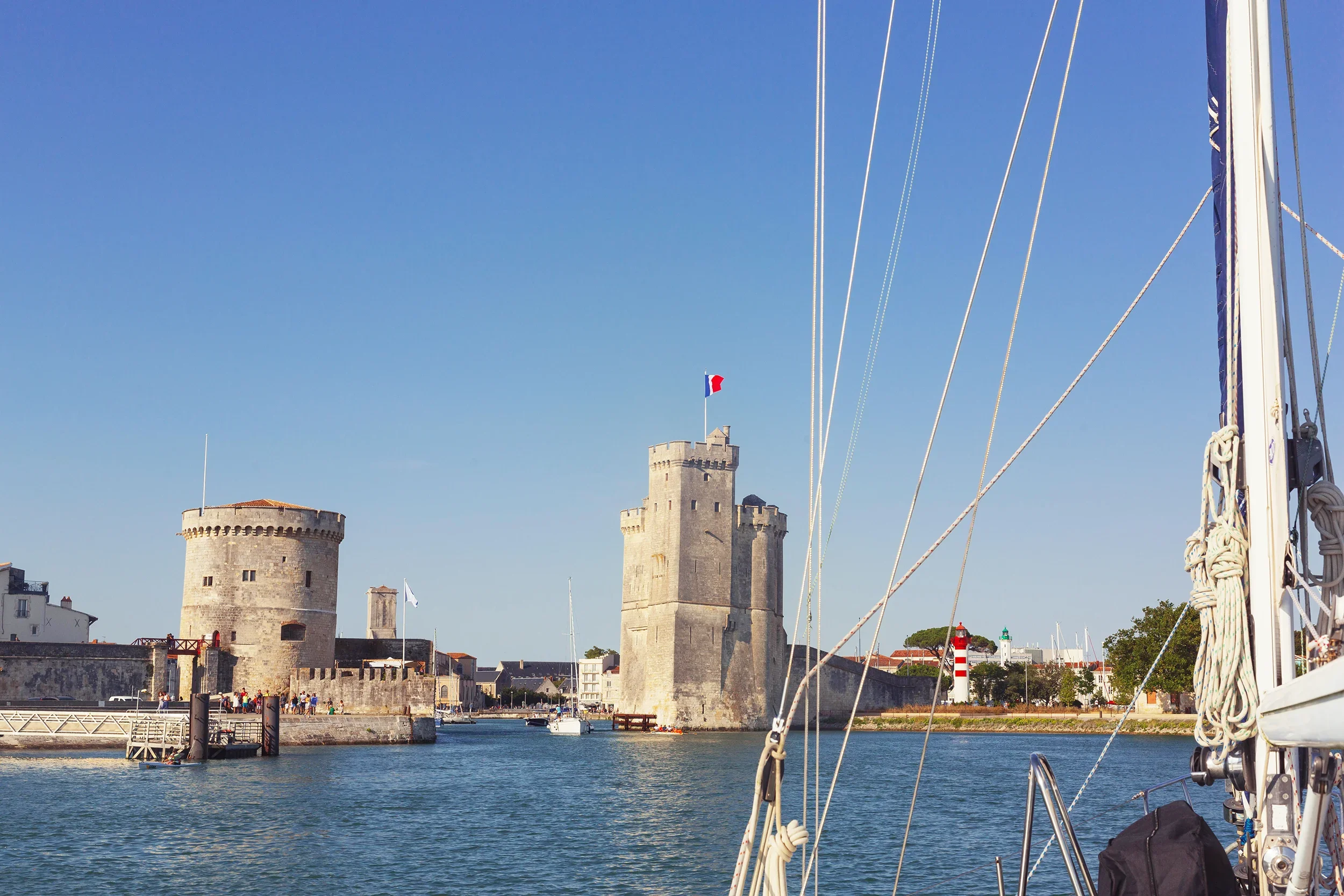 A historic stone castle with a French flag on top, located along a river, with a sailboat docking at the castle's base and a clear blue sky overhead.