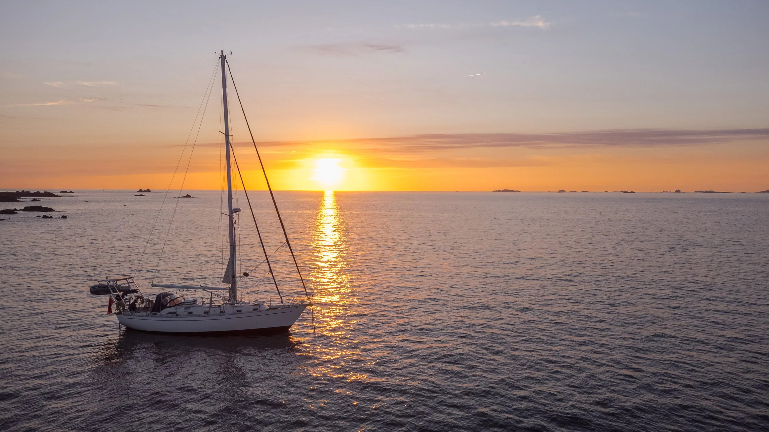A sailboat floating on calm ocean waters during a vibrant sunset with a colorful sky and distant small islands on the horizon.