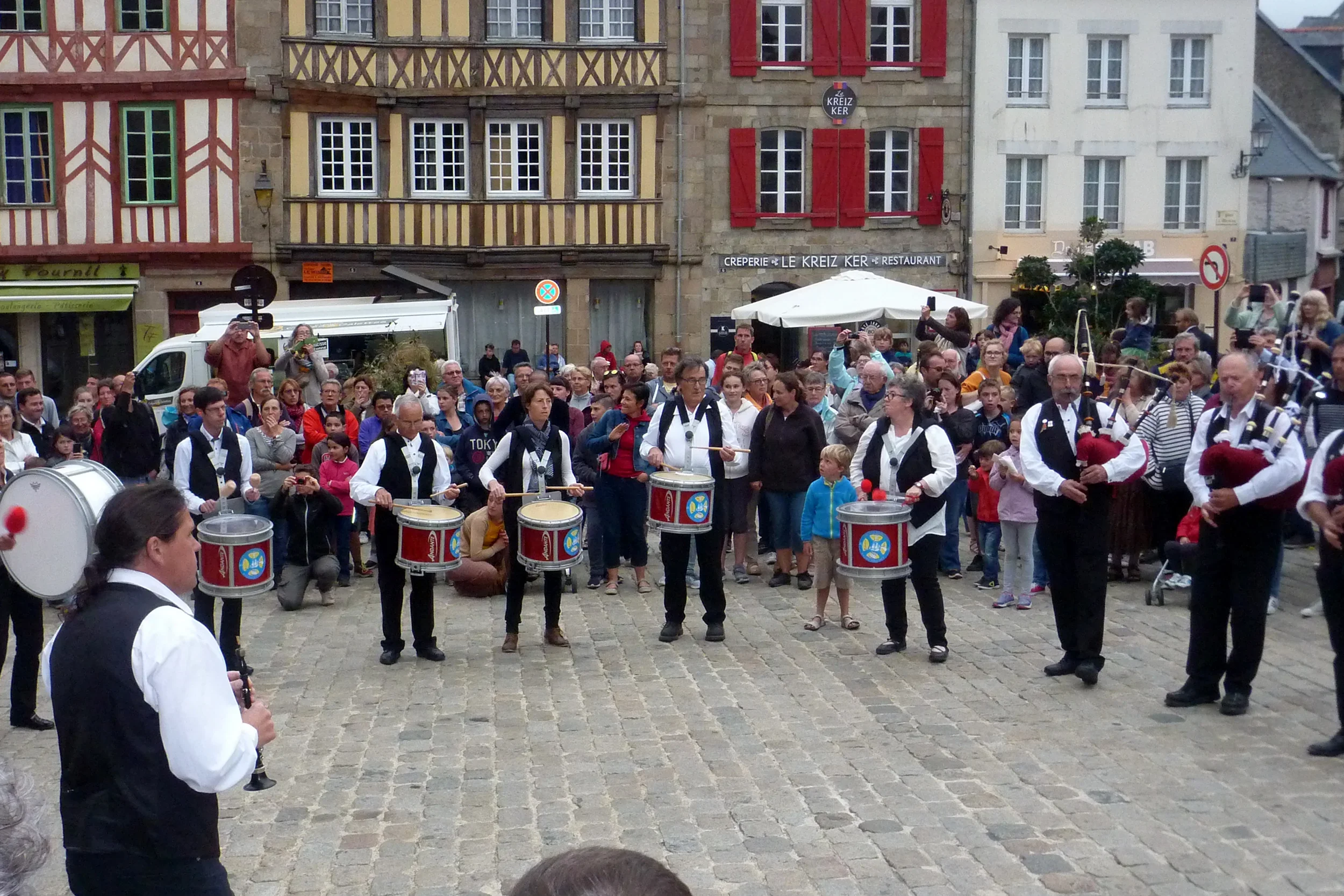 Street performance with a group of musicians playing drums in front of a crowd in a European town square with colorful old buildings.