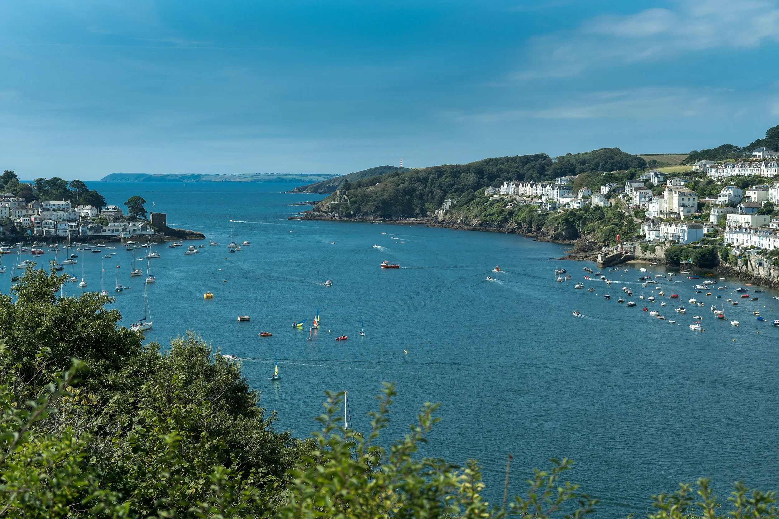 A coastal town with white houses on a hillside overlooking a harbor filled with sailboats and small boats, with lush green trees and rolling hills in the background.