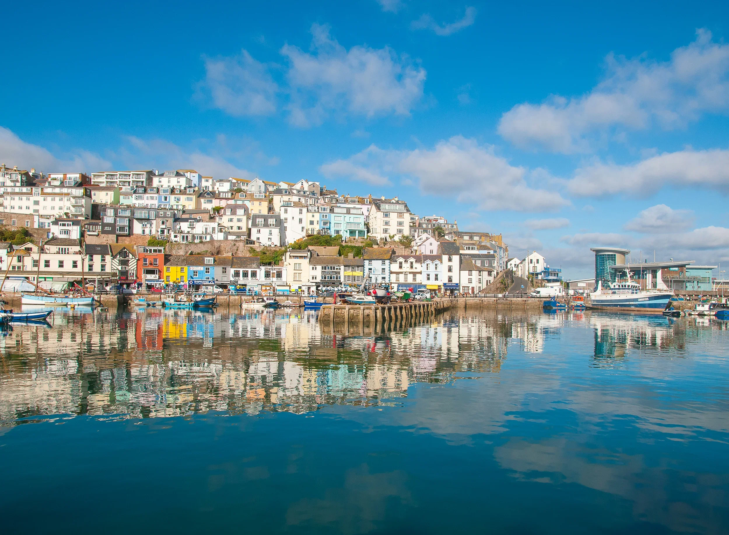 Colorful houses and boats reflecting in a calm harbor under a partly cloudy blue sky.