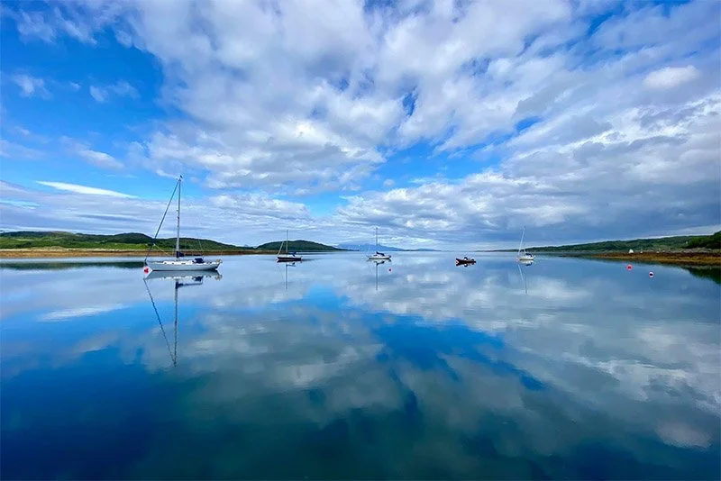 Calm water with sailboats and a small boat, with a partly cloudy sky reflected on the surface, and green hills on the horizon.