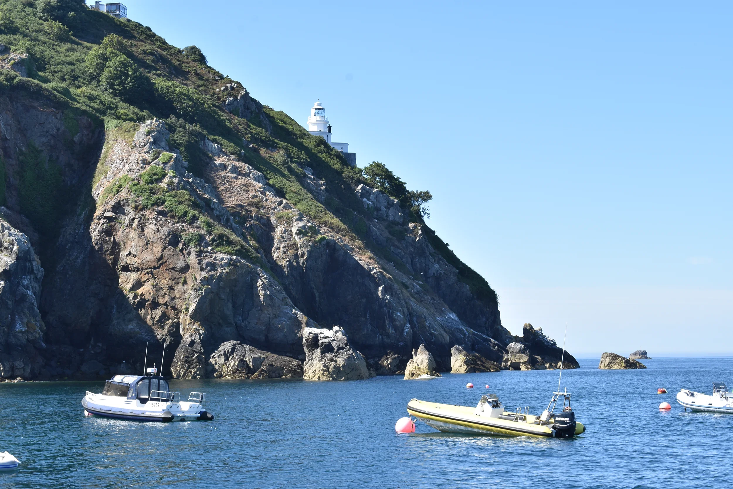 Boats anchored in calm water near a rocky, green hillside with a white lighthouse at the top, under a clear blue sky.