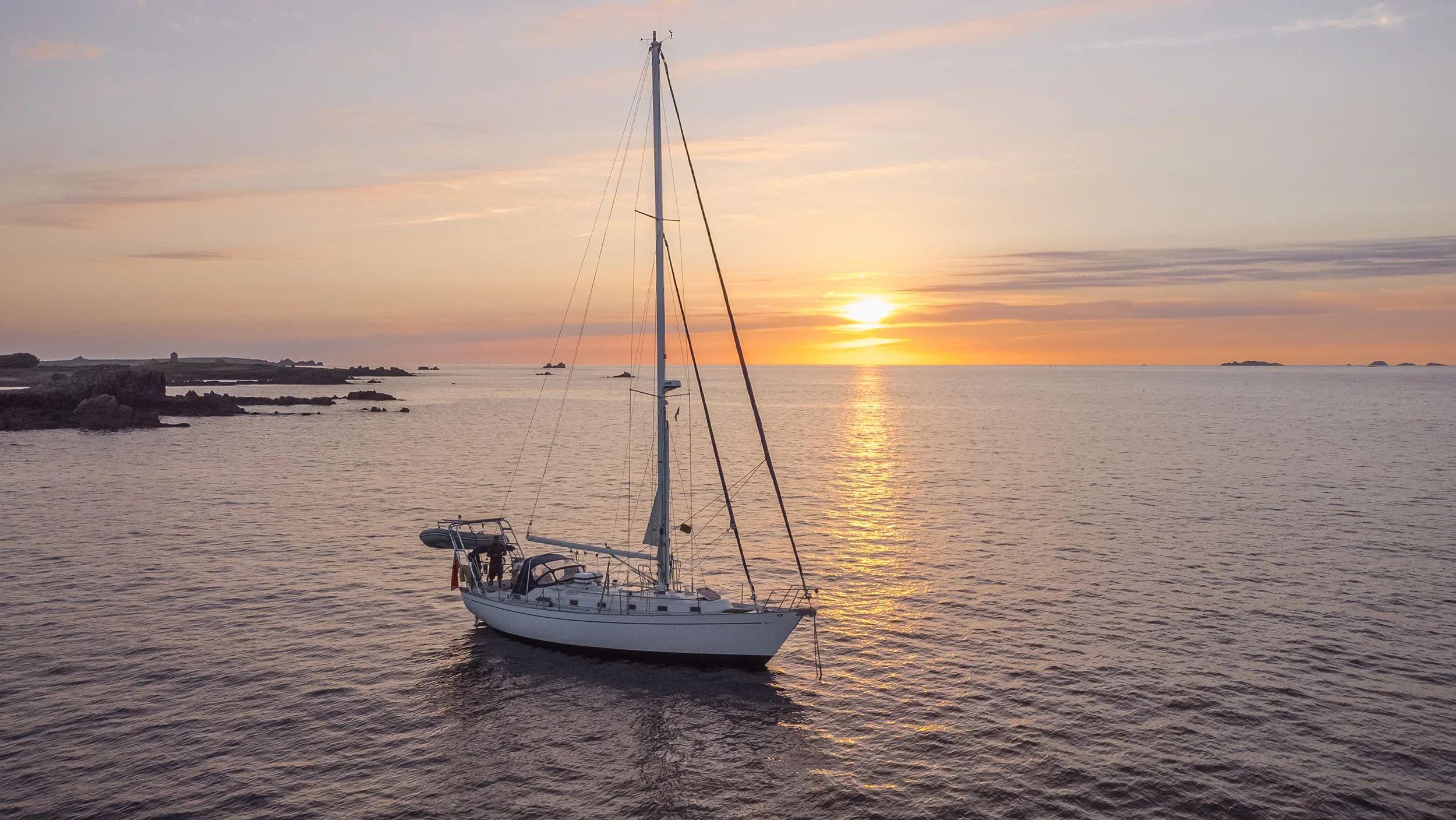 A sailboat floating on calm water during a sunset, with the sky painted in soft pastel colors and a few small islands or rocks in the distance.