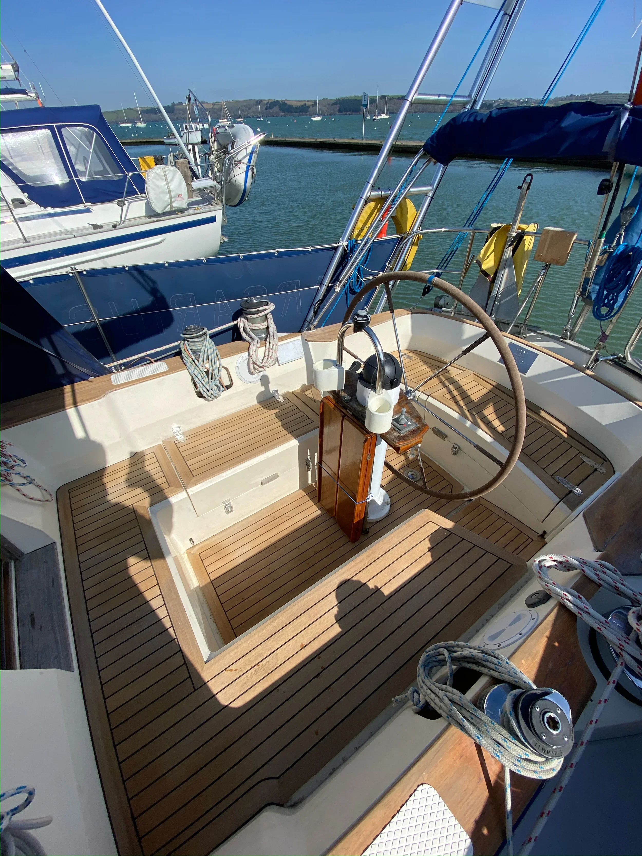 View of a sailboat's cockpit with a wooden floor, steering wheel, and various ropes, docked at a marina with other boats and water in the background.