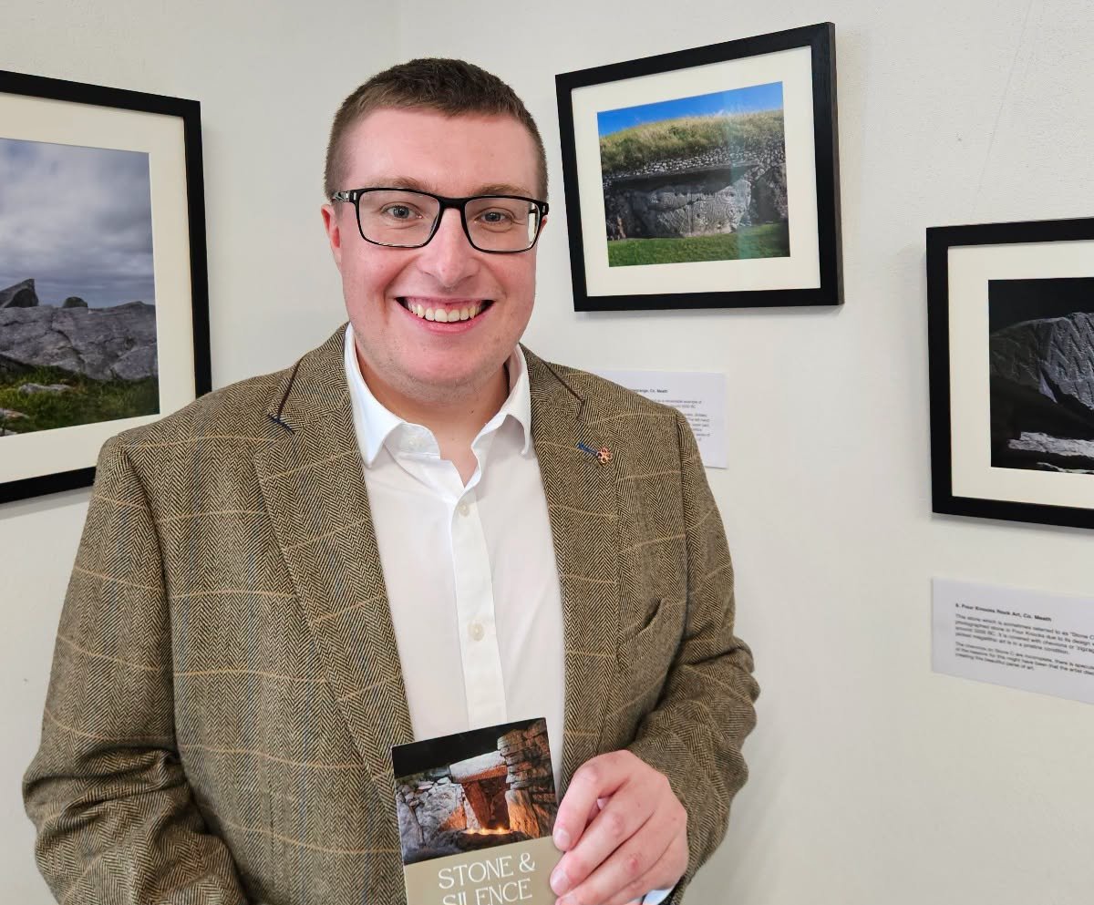 Artist Corey Davenport standing in a brown tweed blazer and glasses holding a pamphlet at an art exhibit Called Stone & Silence  with landscape photographs framed and hung on a white wall.