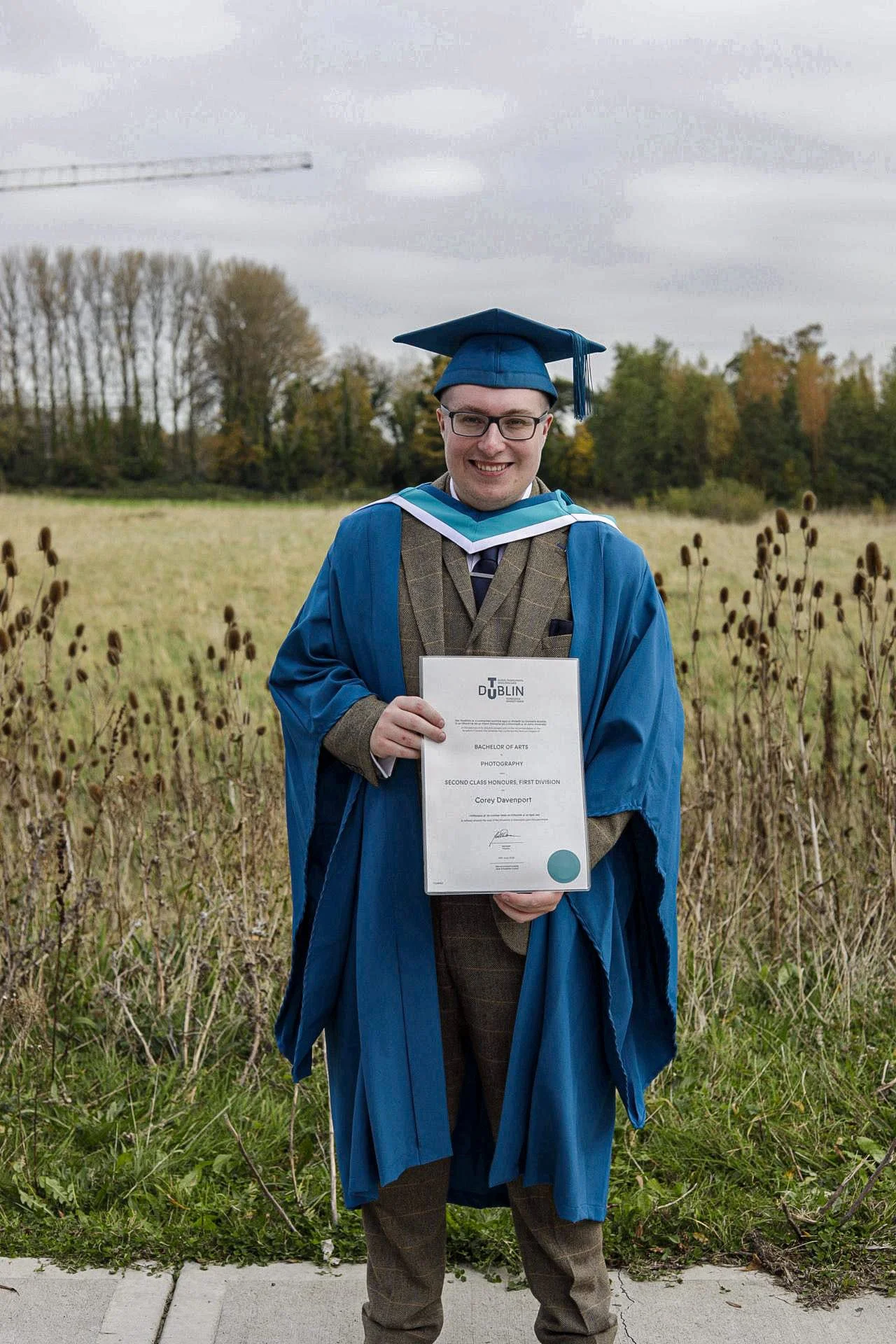 A person in a graduation cap and gown holding a diploma outdoors in a field with trees in the background.