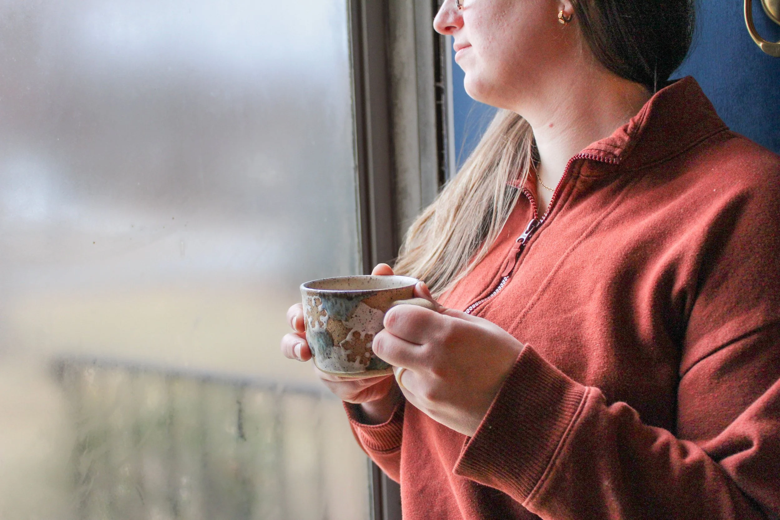 A woman holding a ceramic mug and looking out a window, dressed in a red-orange zip-up sweater.
