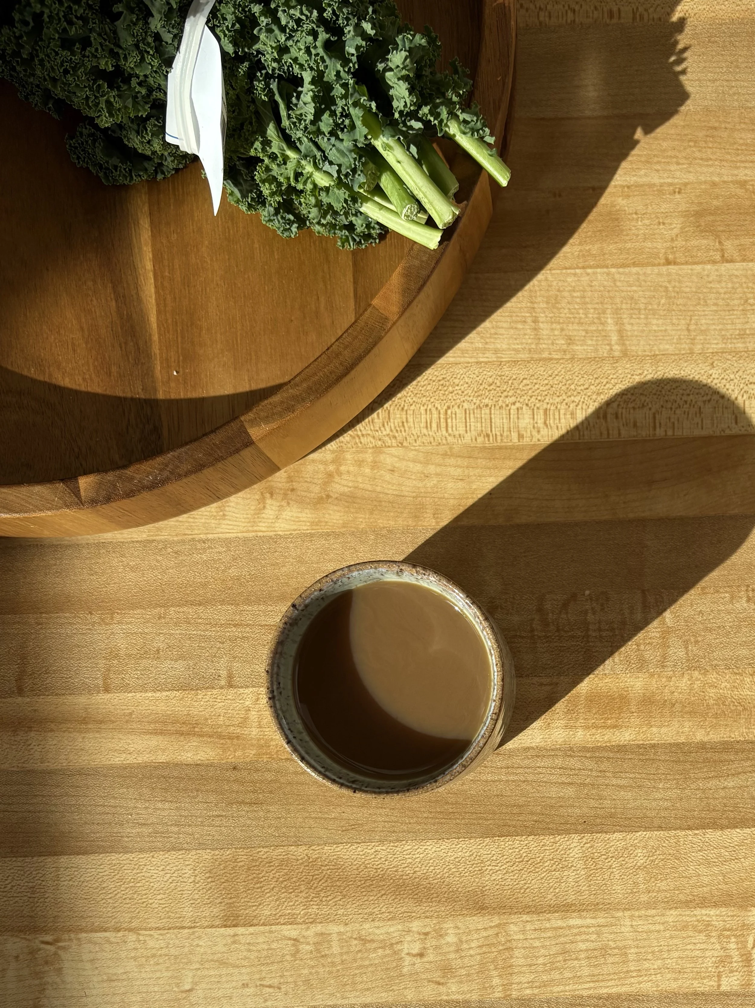 A wooden table with a cup of coffee and a bunch of kale on a cutting board.