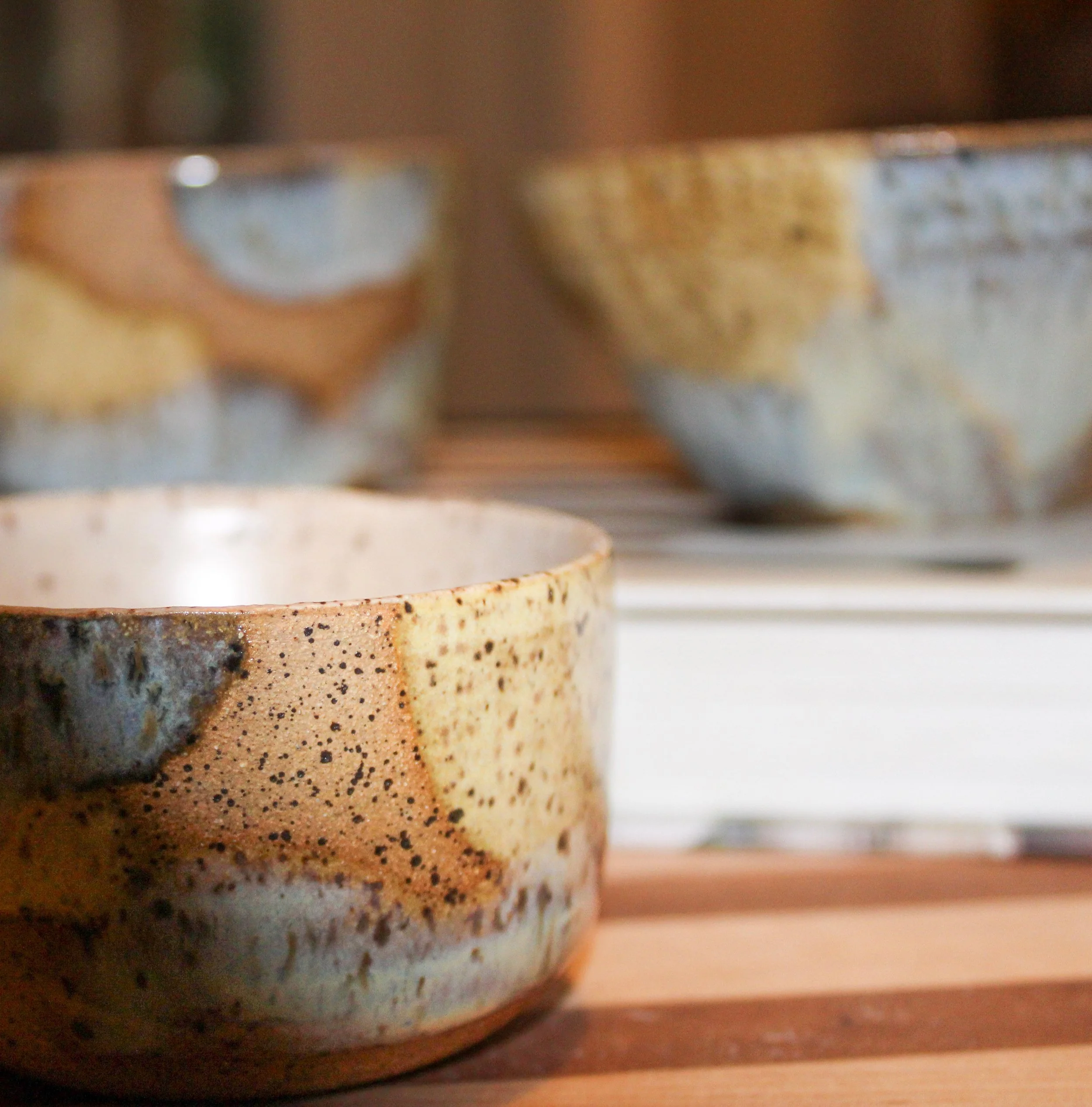 Close-up of a ceramic bowl with a speckled glaze, with two more blurry bowls in the background on a wooden surface.