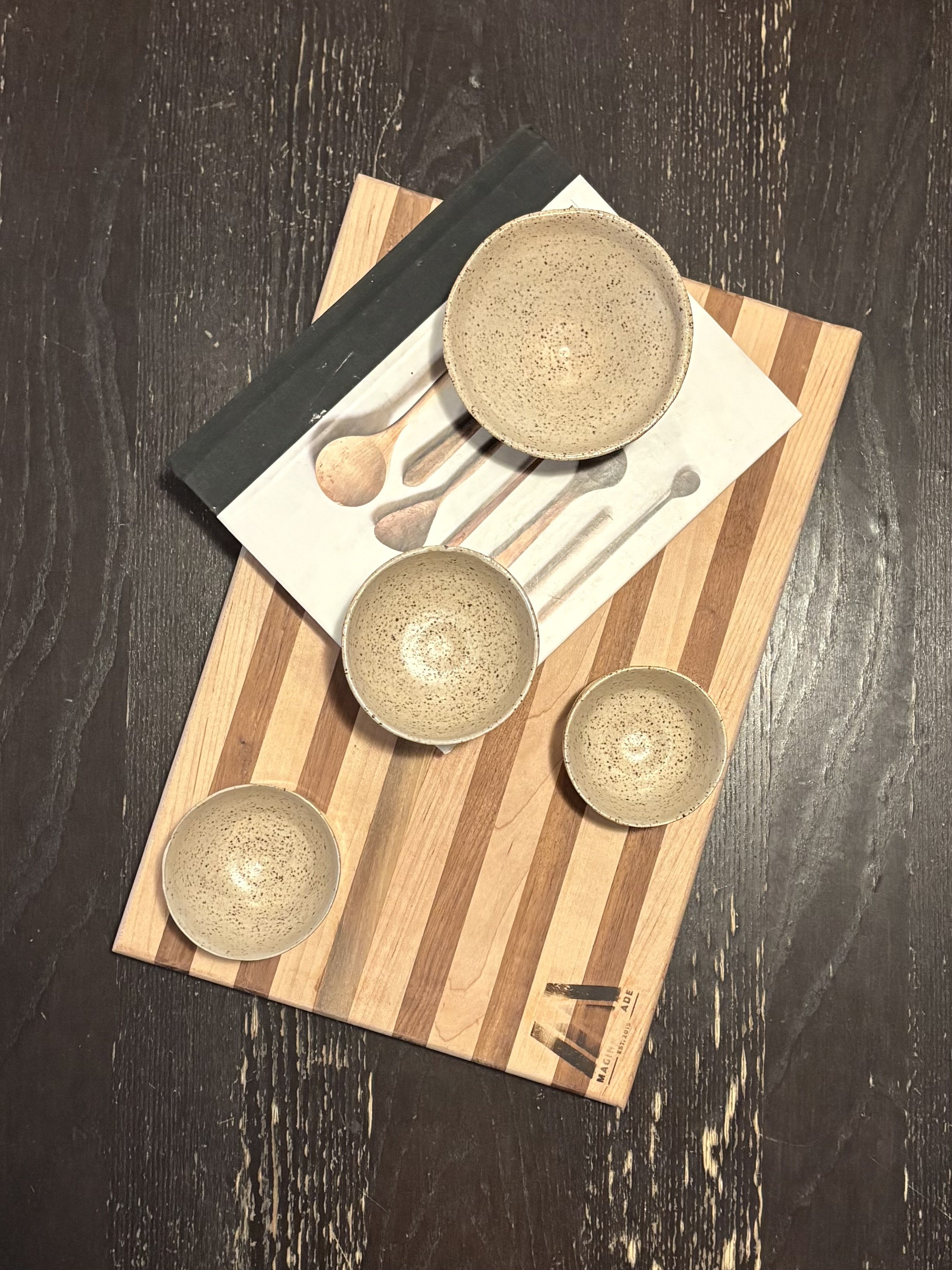 A wooden serving board with four small speckled beige bowls, a white book with images of spoons, and a small beige bowl on top, all on a dark wooden table.