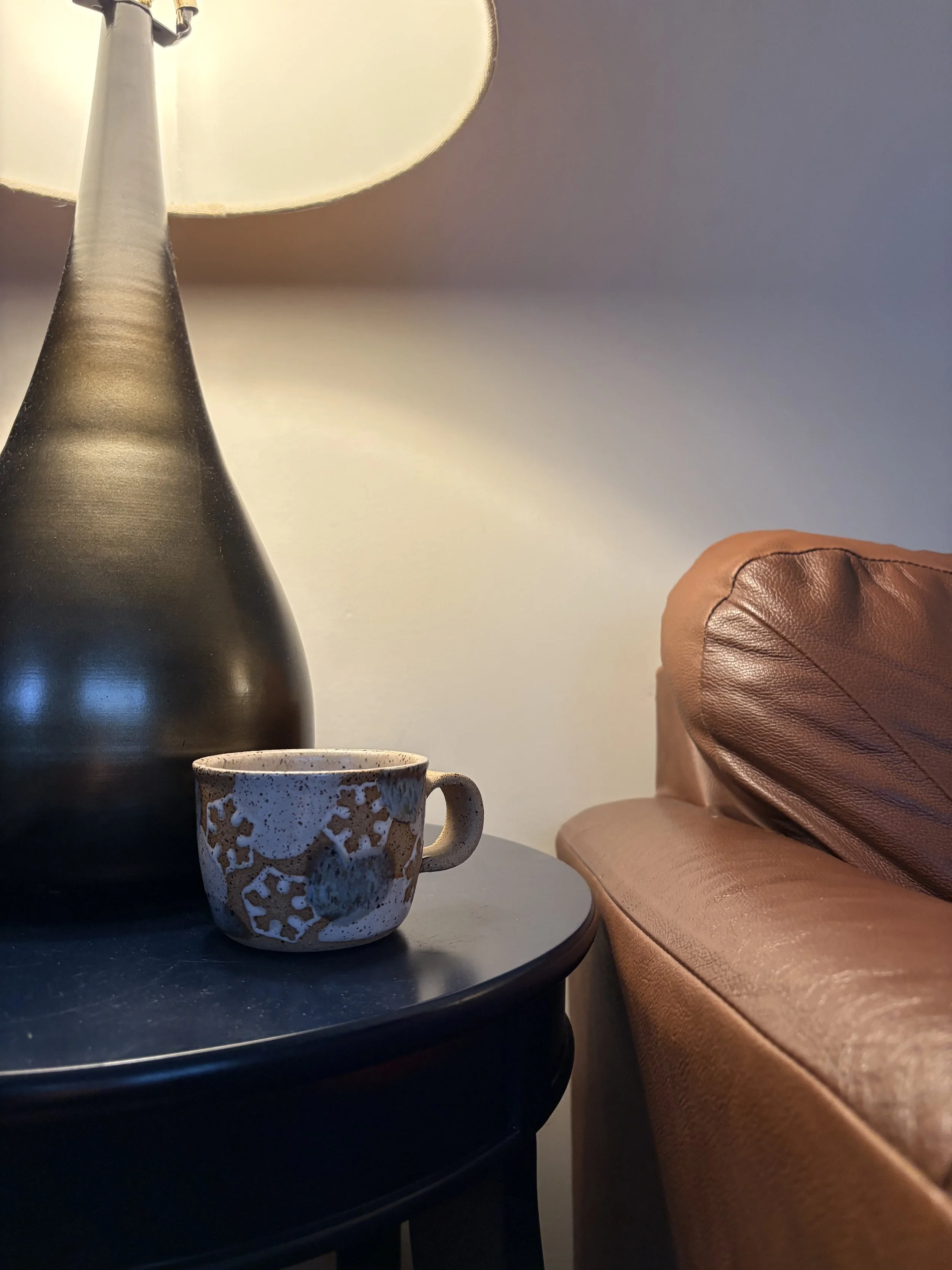 A black side table with a ceramic mug featuring snowflake designs, a large black lamp with a cream lampshade, and a brown leather couch to the right, against a plain light-colored wall.