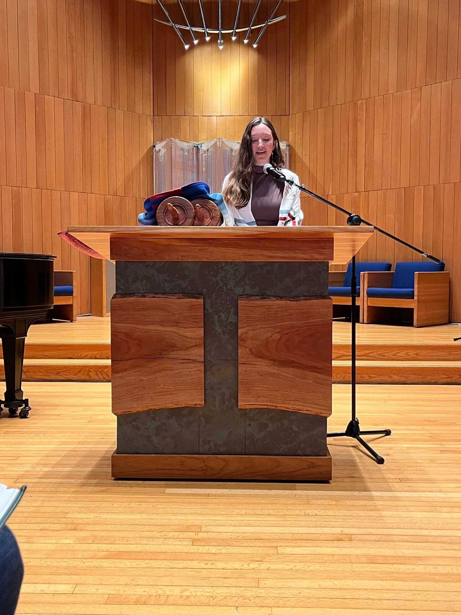 A woman stands at a wooden podium with a microphone in a wood-paneled room, possibly a church or auditorium, reading or speaking. There are chairs with blue cushions behind her and some fabric or cloth on the podium.
