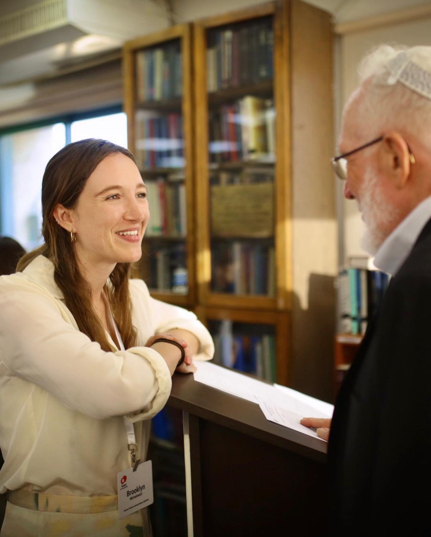 A woman with a name tag that says Brooklyn is smiling and talking to a man with a beard and glasses at a reception desk in a library or bookstore, with bookshelves in the background.