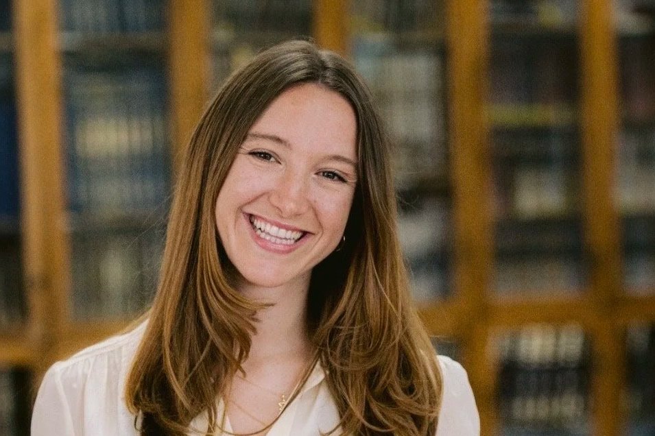 Young woman with long brown hair smiling in front of a bookshelf in a library or study.