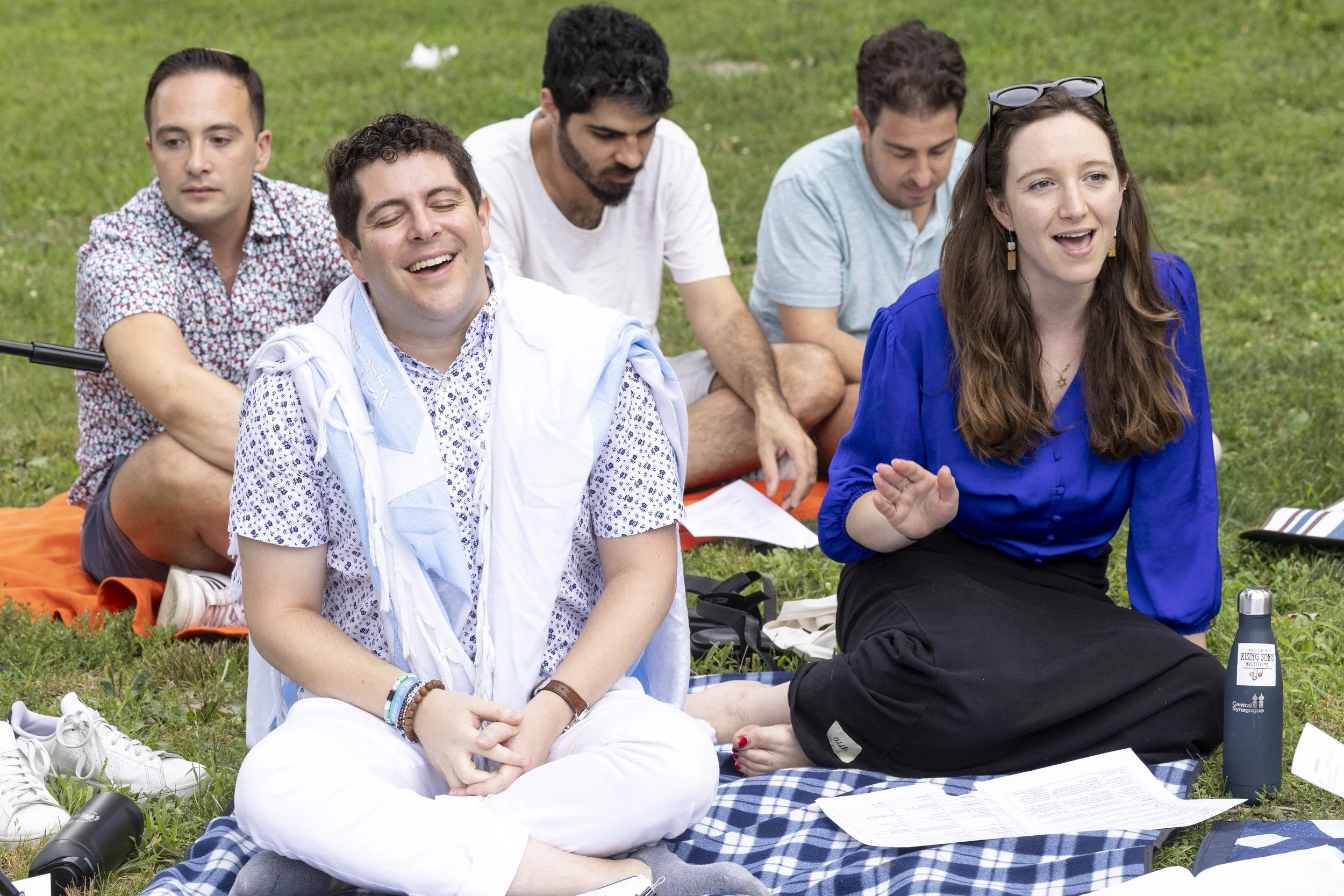 Group of people sitting on grass at an outdoor gathering, with some smiling and engaging in conversation.