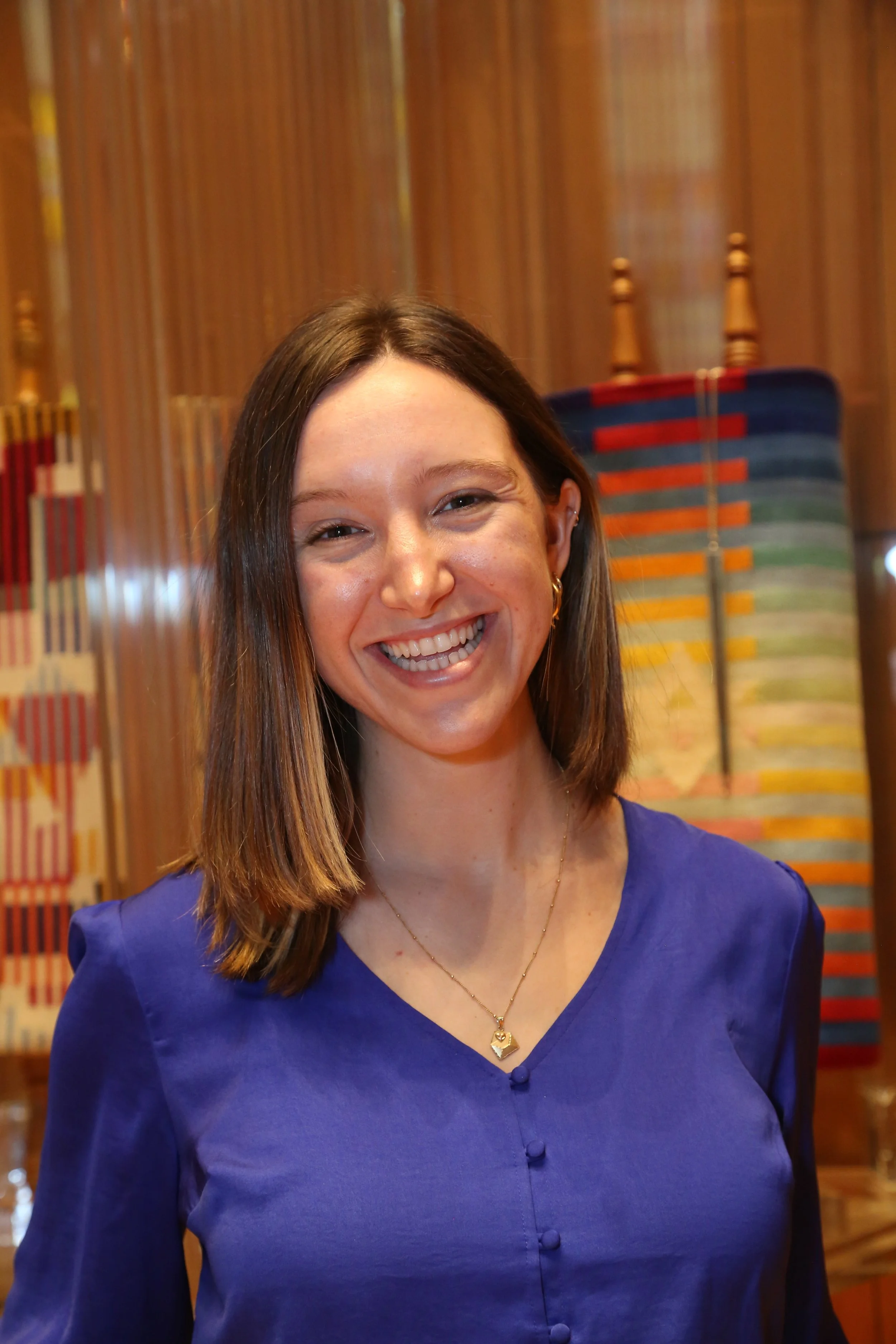 Happy woman with shoulder-length brown hair, wearing a royal blue top and a gold necklace, smiling at the camera in a warmly decorated indoor setting.