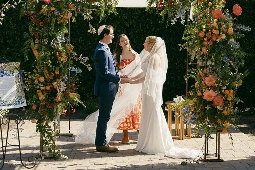 A wedding ceremony with a bride and groom holding hands, standing under a floral arch, with a officiant smiling in the background.