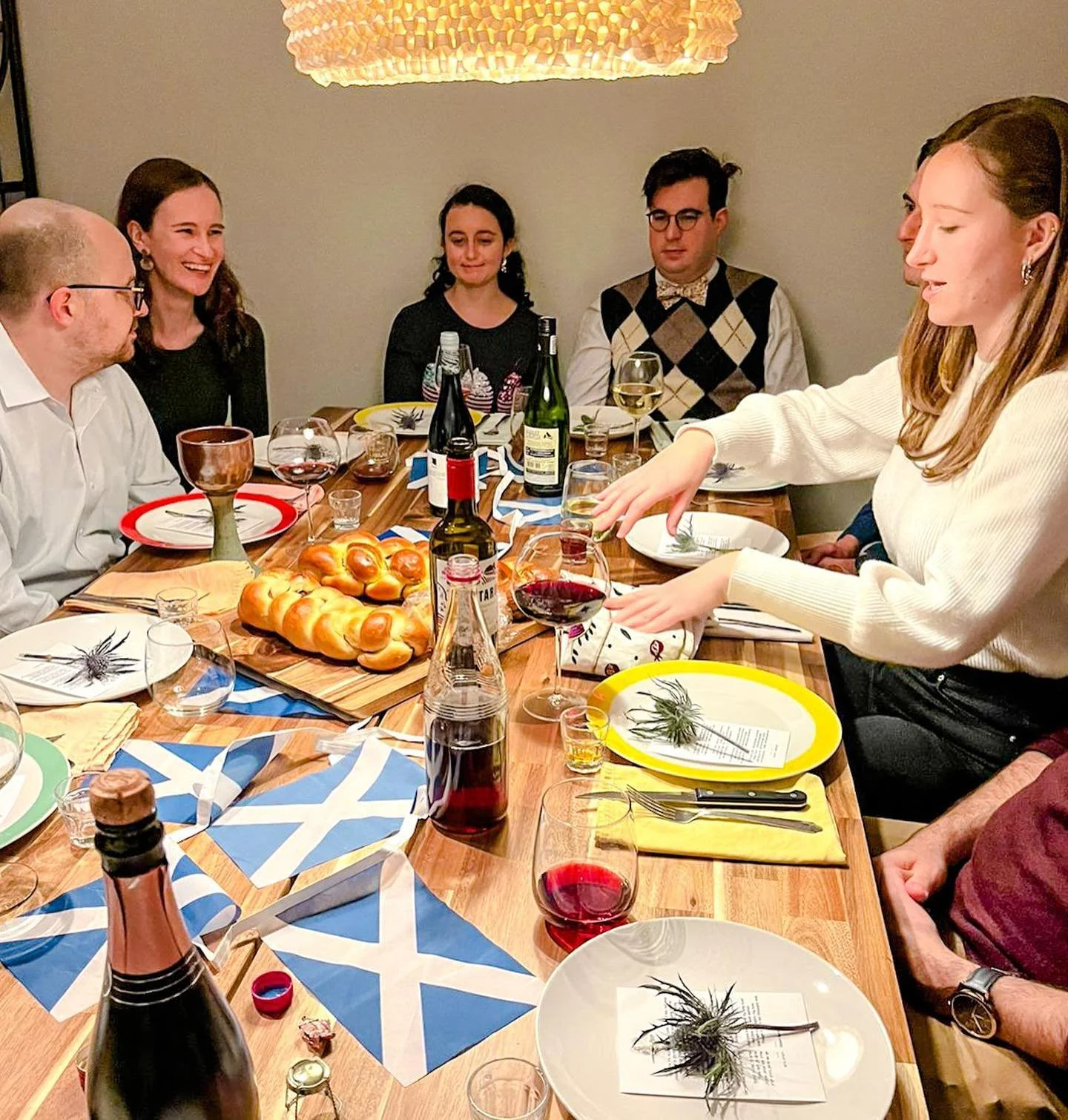 People gathered around a dinner table with food and drinks, preparing to celebrate, possibly a festive occasion with themed decorations and bread in the center.