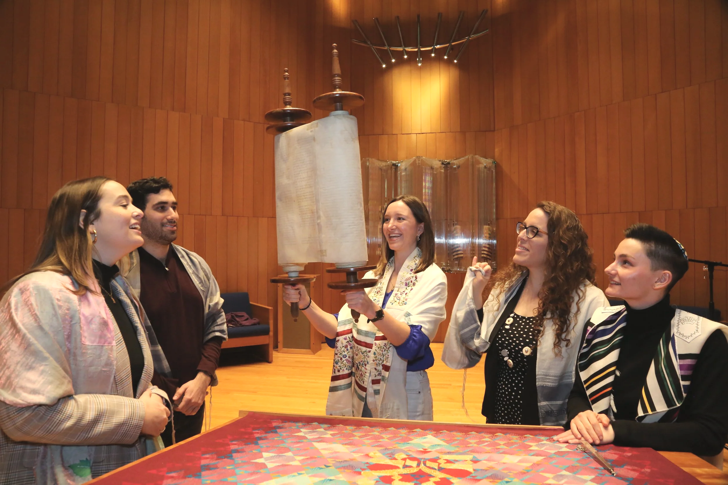 A woman holding two torah scrolls standing in front of four people in a wooden room, engaging in a celebration or ceremony.
