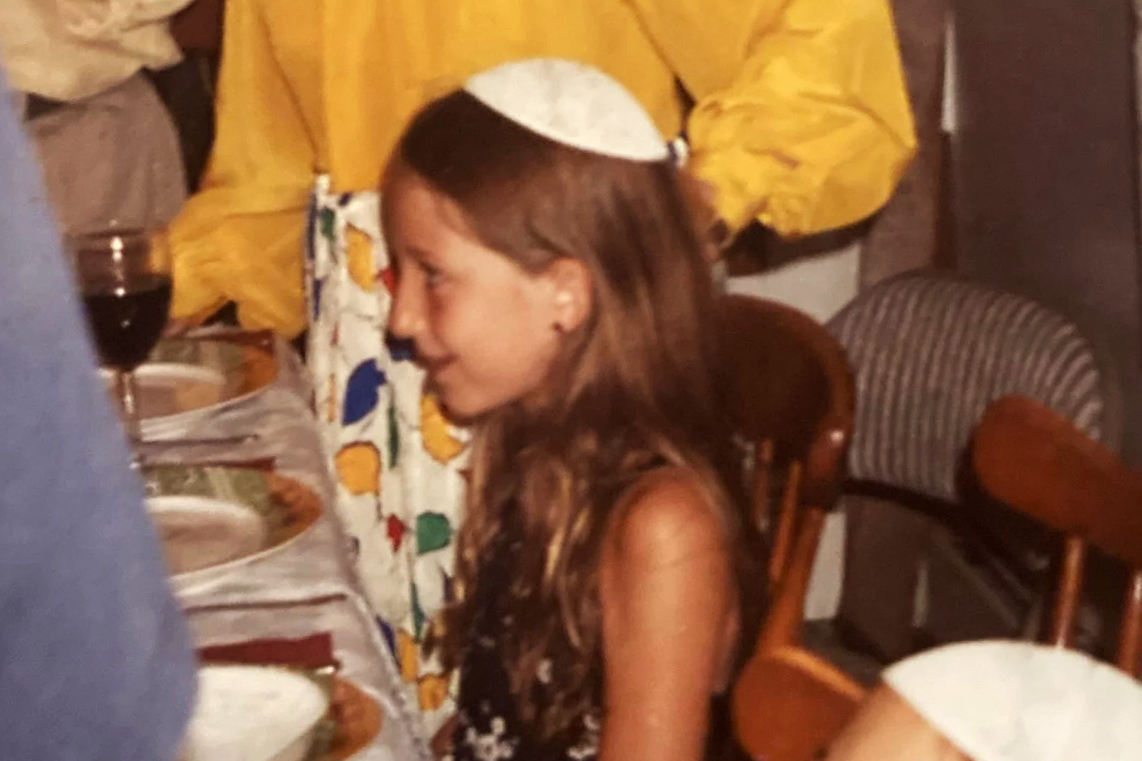 A young girl with long brown hair, wearing a white kippah, is sitting at a table during a dinner or celebration. There are plates, a glass of red wine, and a floral dress worn by someone next to her. The background includes chairs and a person dressed in yellow.