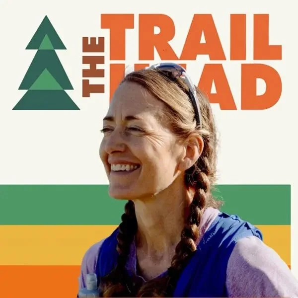 Author and ultrarunner Sarah Lavender Smith with braided hair smiling, wearing sunglasses on her head and a blue shirt, standing in front of a backdrop with a trail logo and green, yellow, and orange stripes.