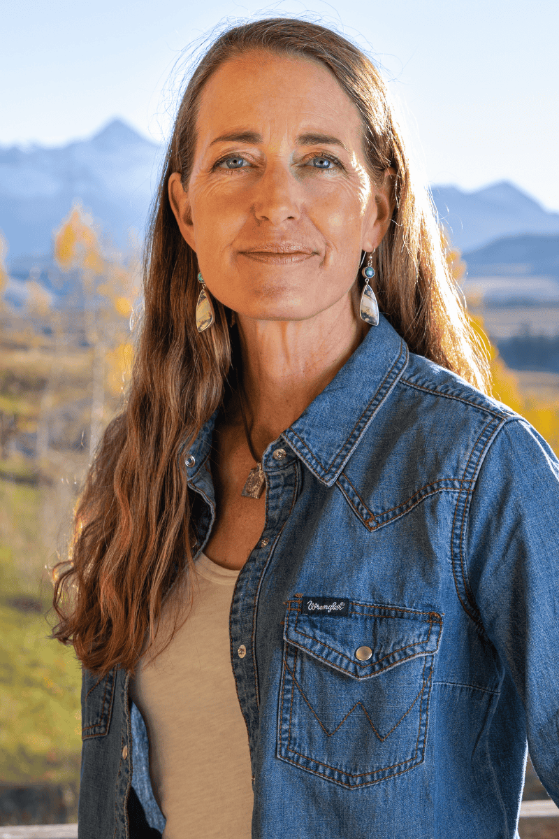 Author and ultrarunner Sarah Lavender Smith with long brown hair, wearing a denim jacket and earrings, standing outdoors in front of mountains and trees.