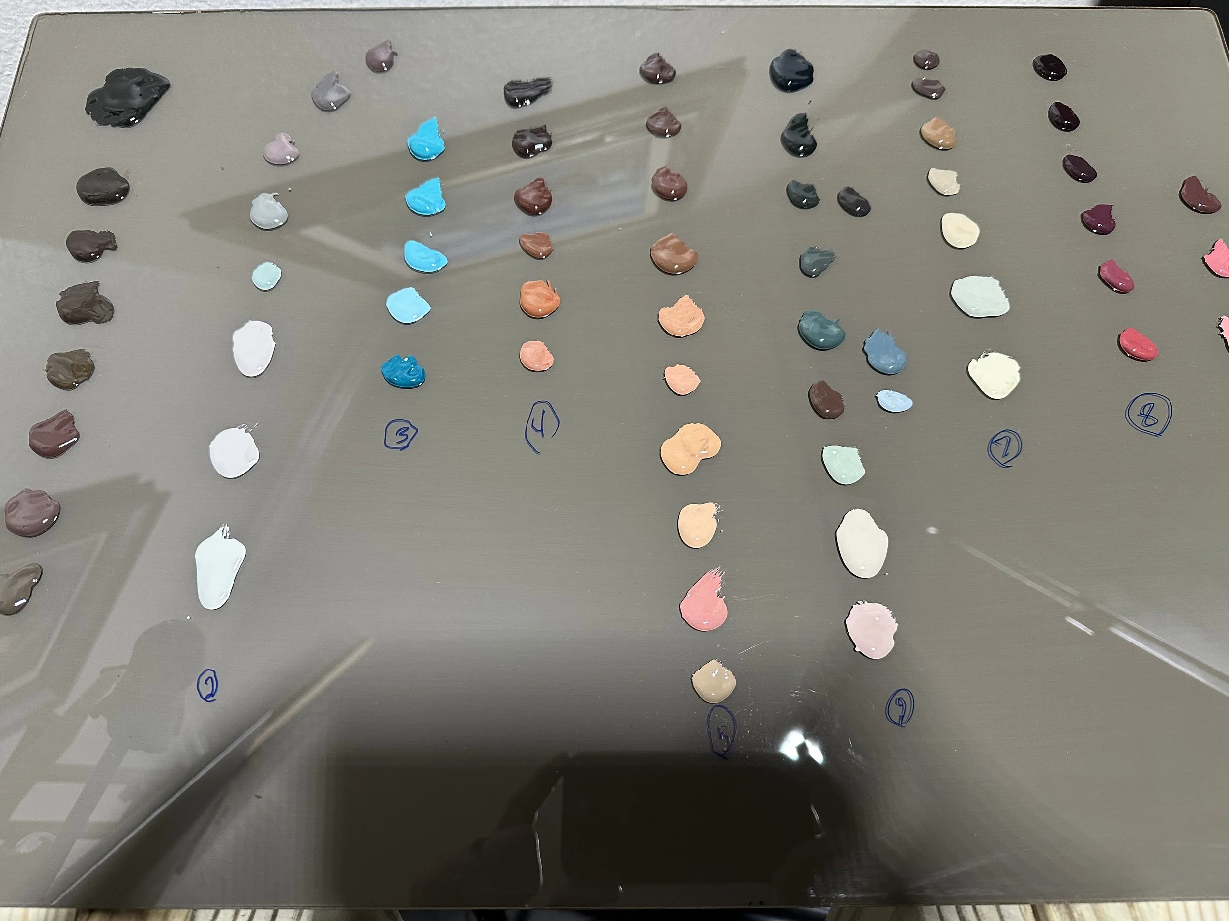 Close-up of a glass artist palette with oil paint colors arranged in numbered rows for color theory mixing. Displays gradients of earth tones, cool blues, and warm skin tones for realistic painting.