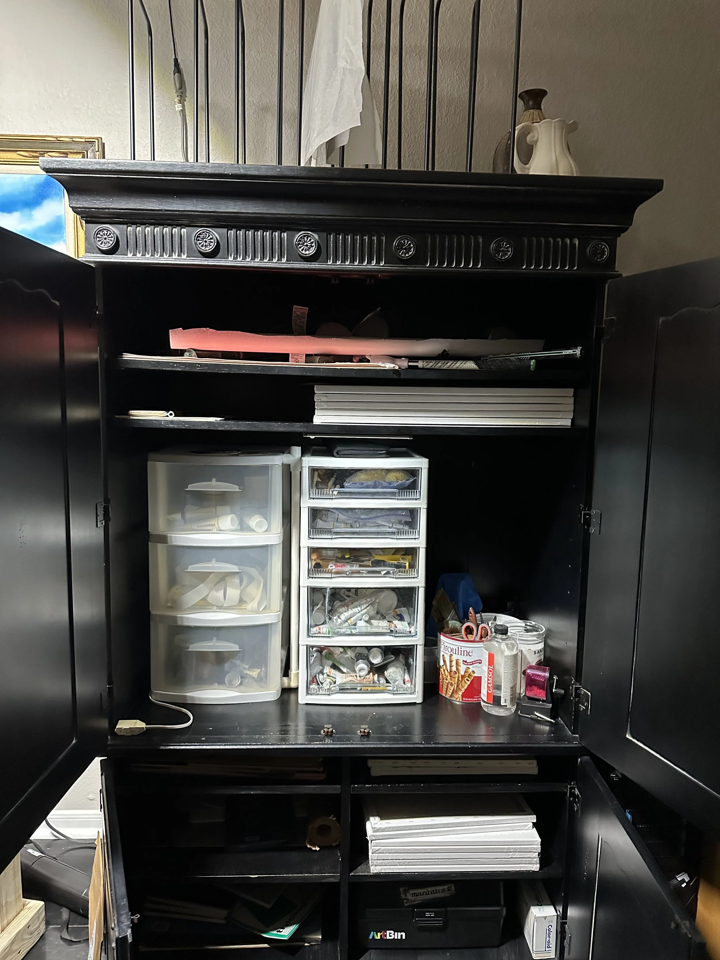 Large distressed black armoire with decorative rosette molding used for art supply storage in Timothy Chappell's painting studio. The cabinet is opened showing oil painting materials.