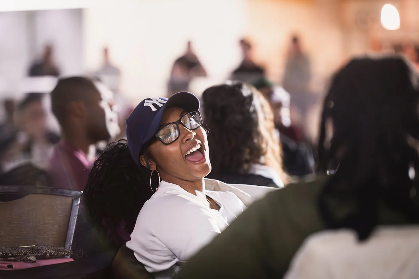 A woman with glasses and a baseball cap laughing during a conference or seminar.