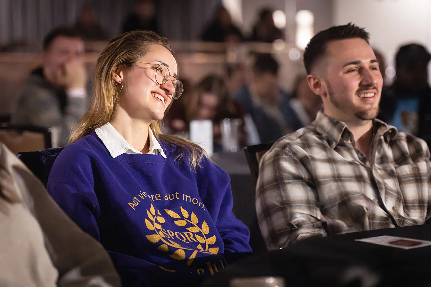 A woman with glasses and long blonde hair laughing, wearing a purple sweatshirt and a white collared shirt beneath, sitting next to a man in a plaid shirt, both attending an indoor event with other people visible in the background.