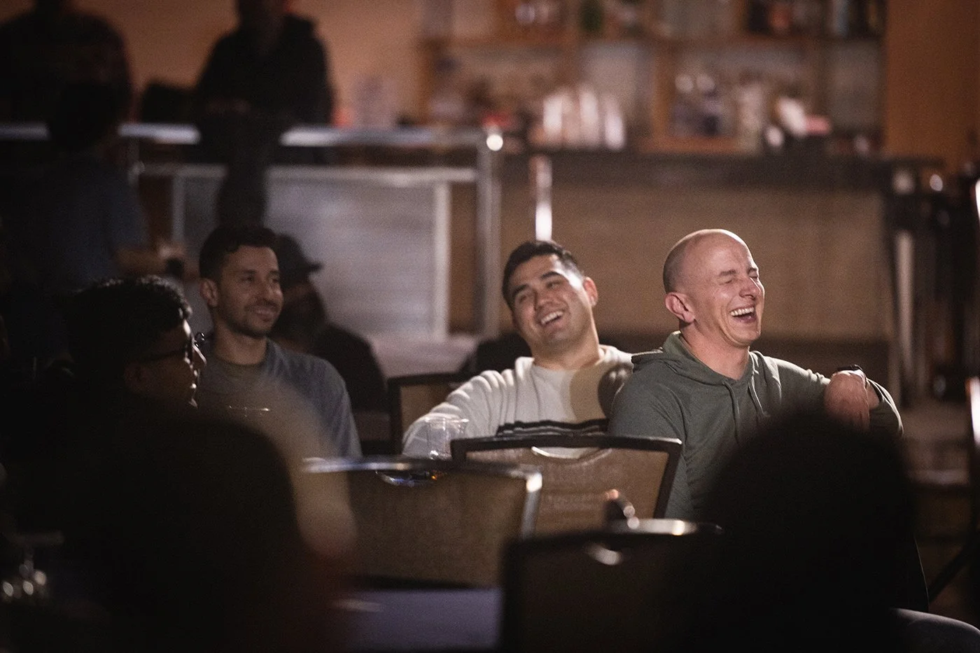 A group of people seated in a dark venue, possibly a comedy club or theater, watching a performance and laughing.