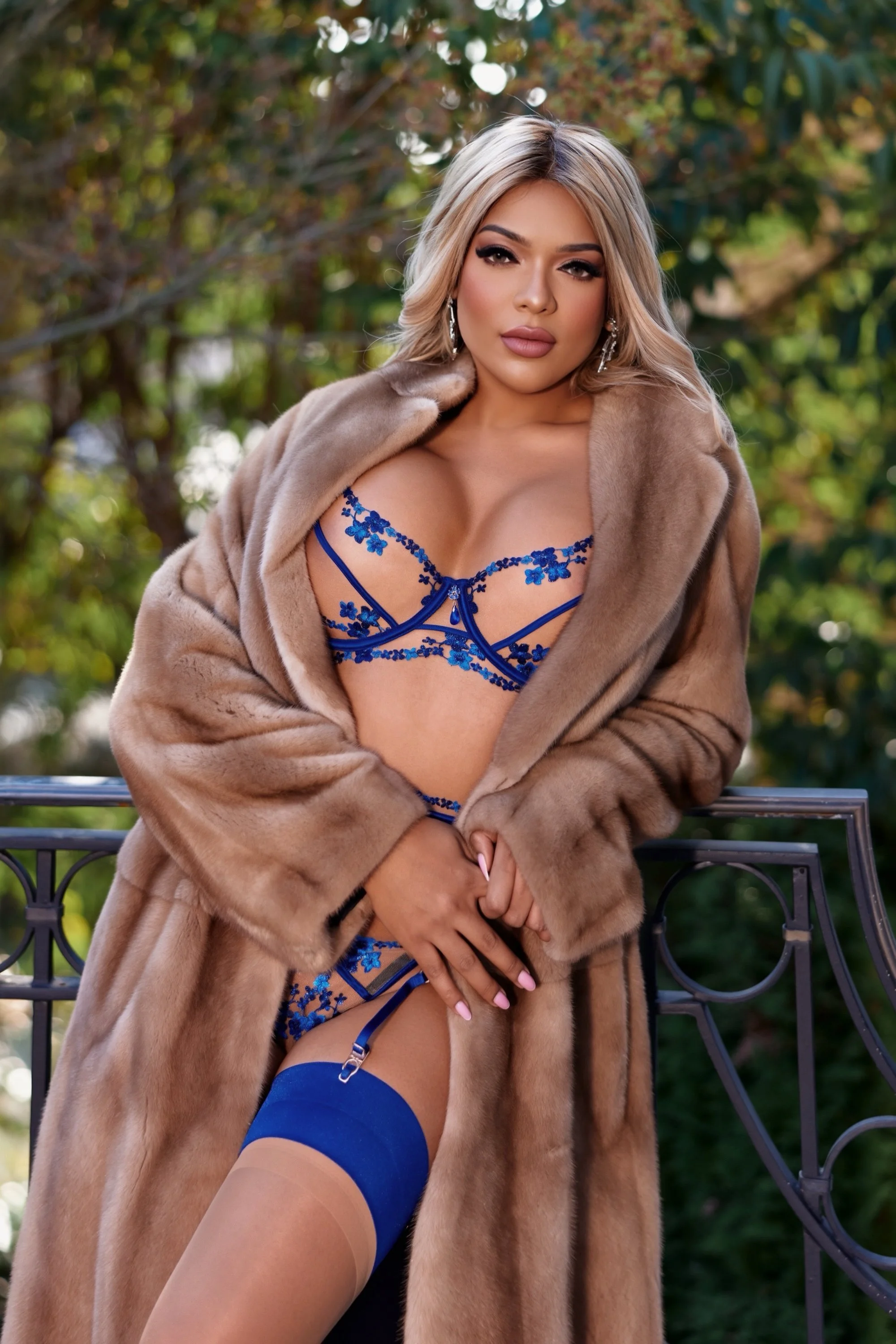 A woman in a fur coat and lingerie, standing outdoors on a balcony with green foliage in the background.