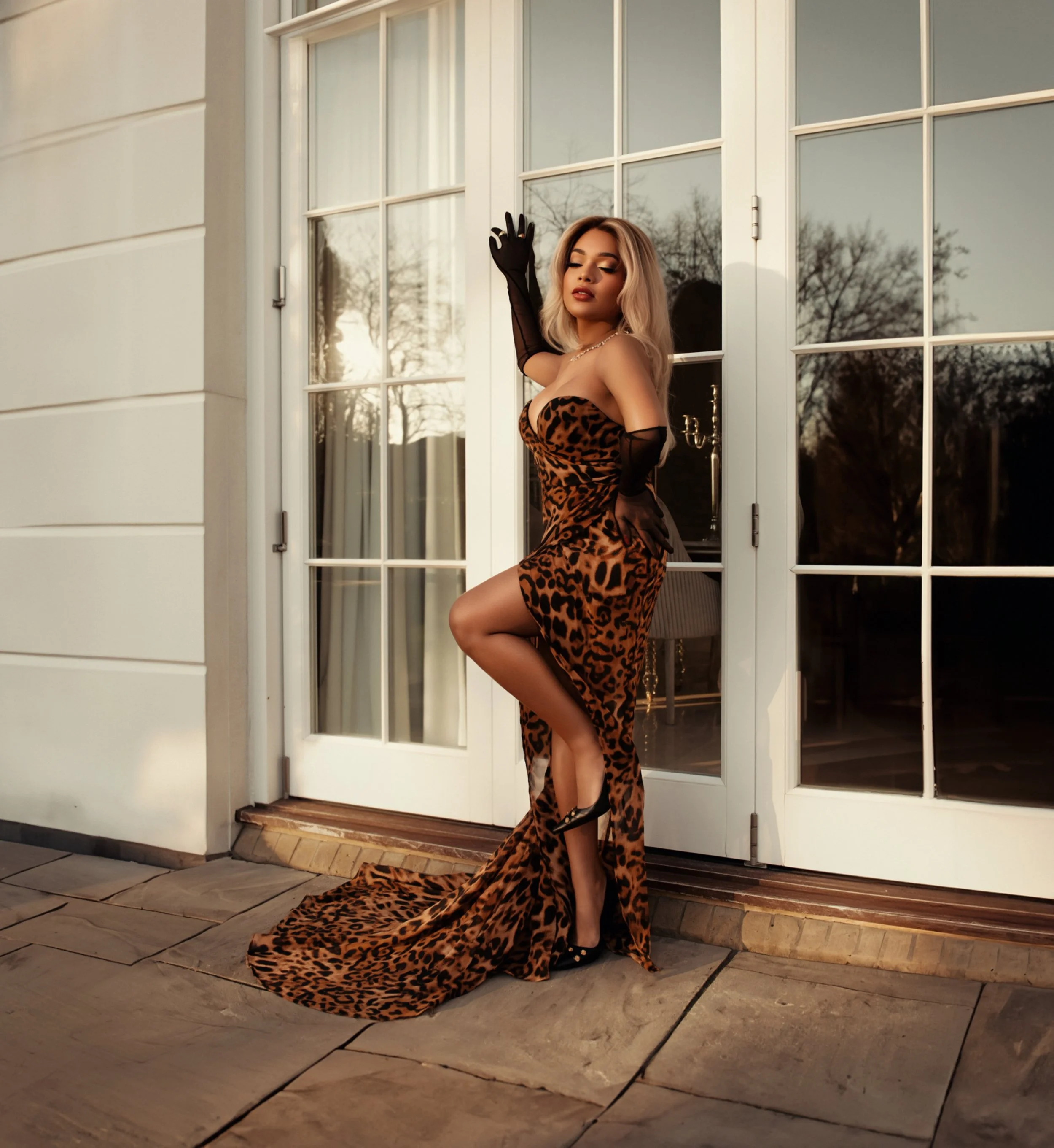 A woman with blonde hair, dressed in a leopard print gown, wearing black gloves and high heels, leaning against a large glass door at sunset.