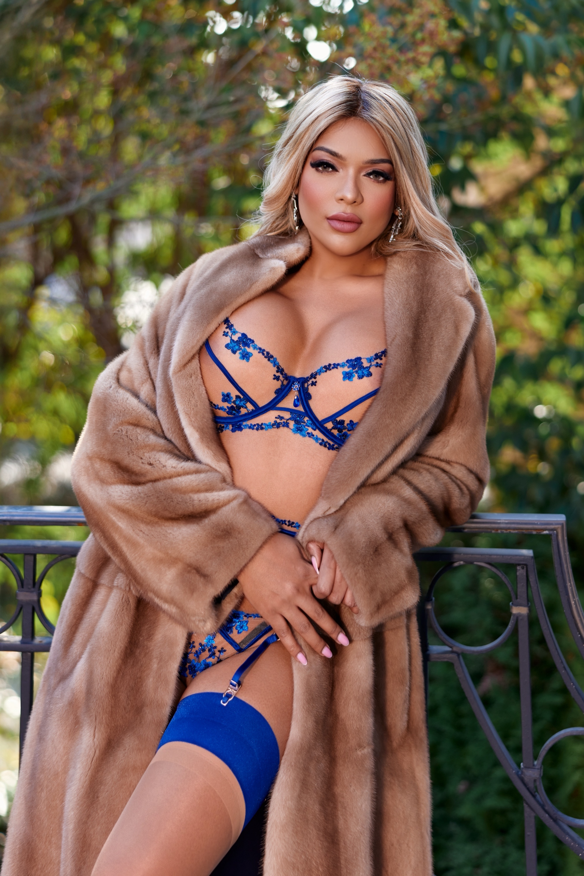 A woman wearing blue lingerie and a fur coat, standing outdoors with trees in the background.