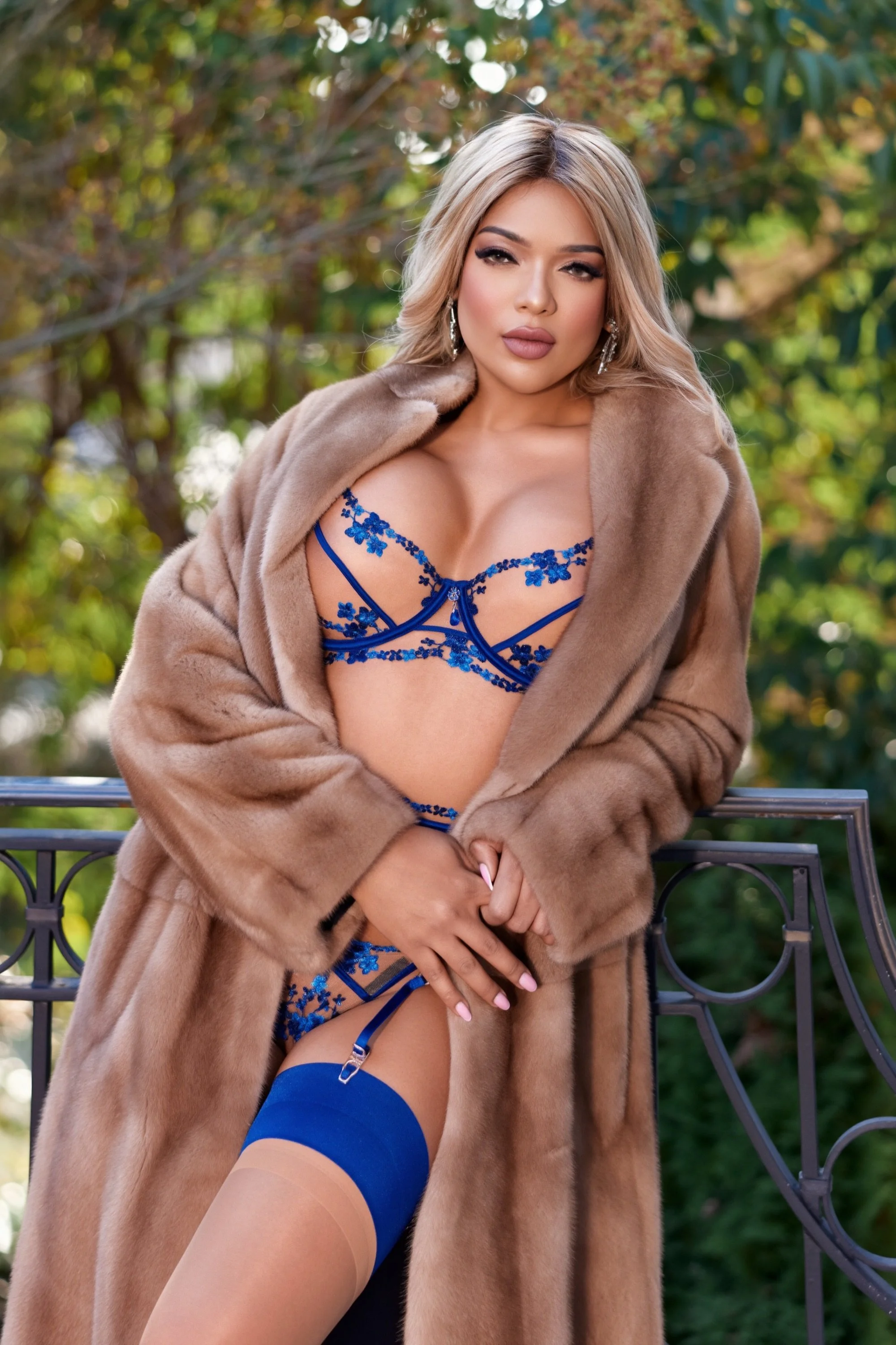 A woman wearing blue lingerie and a beige fur coat stands outdoors on a balcony with a blurred green and yellow foliage background.