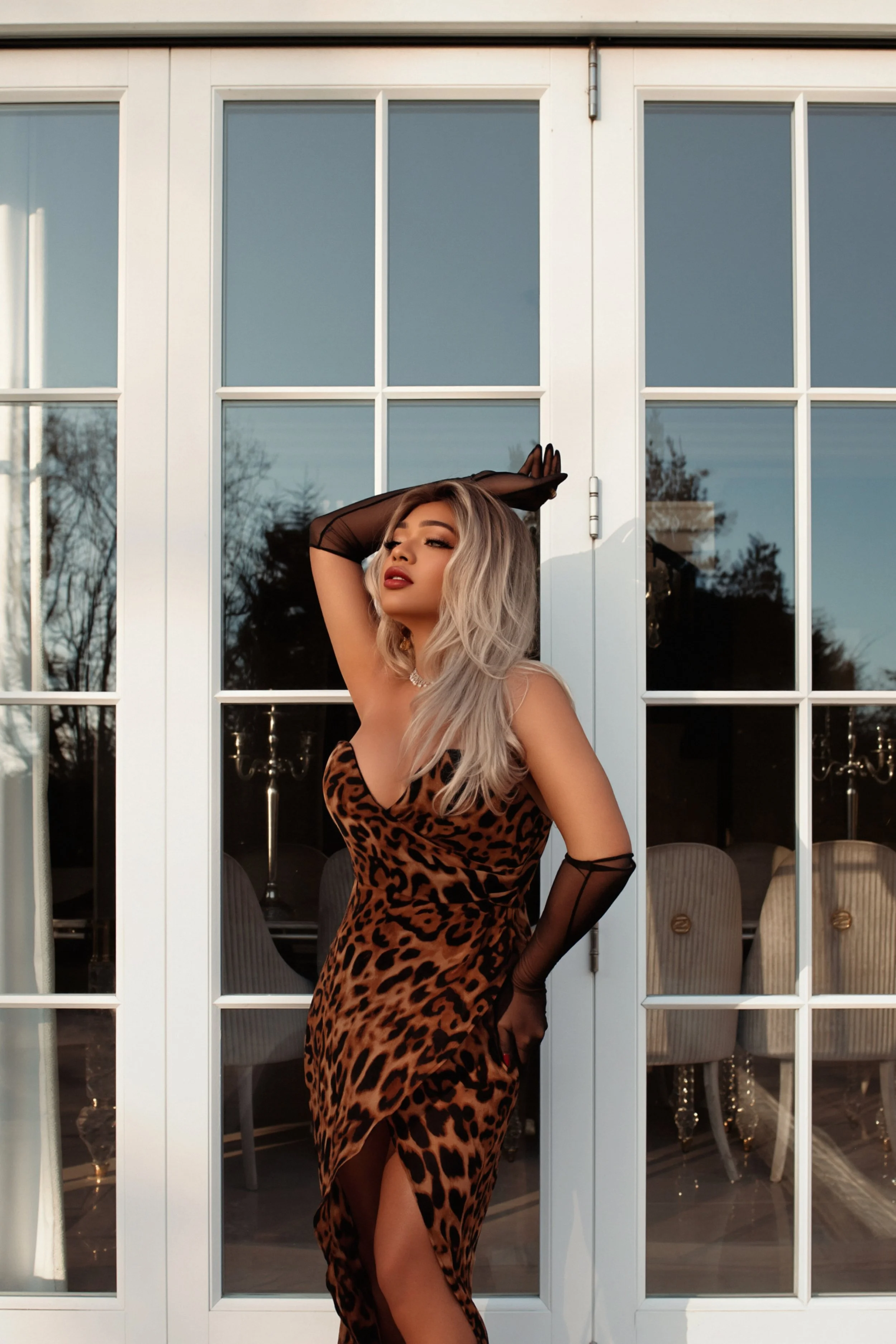 A woman in a leopard print dress with sheer black gloves and blonde hair posing in front of glass doors with reflections of trees and a chandelier inside.